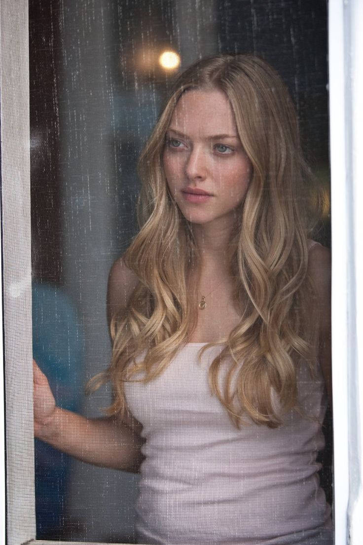 Amanda Seyfried 桌布 壁紙 — 一位長金髮的女性在夜晚透過雨痕斑斑的窗戶沉思地凝視。