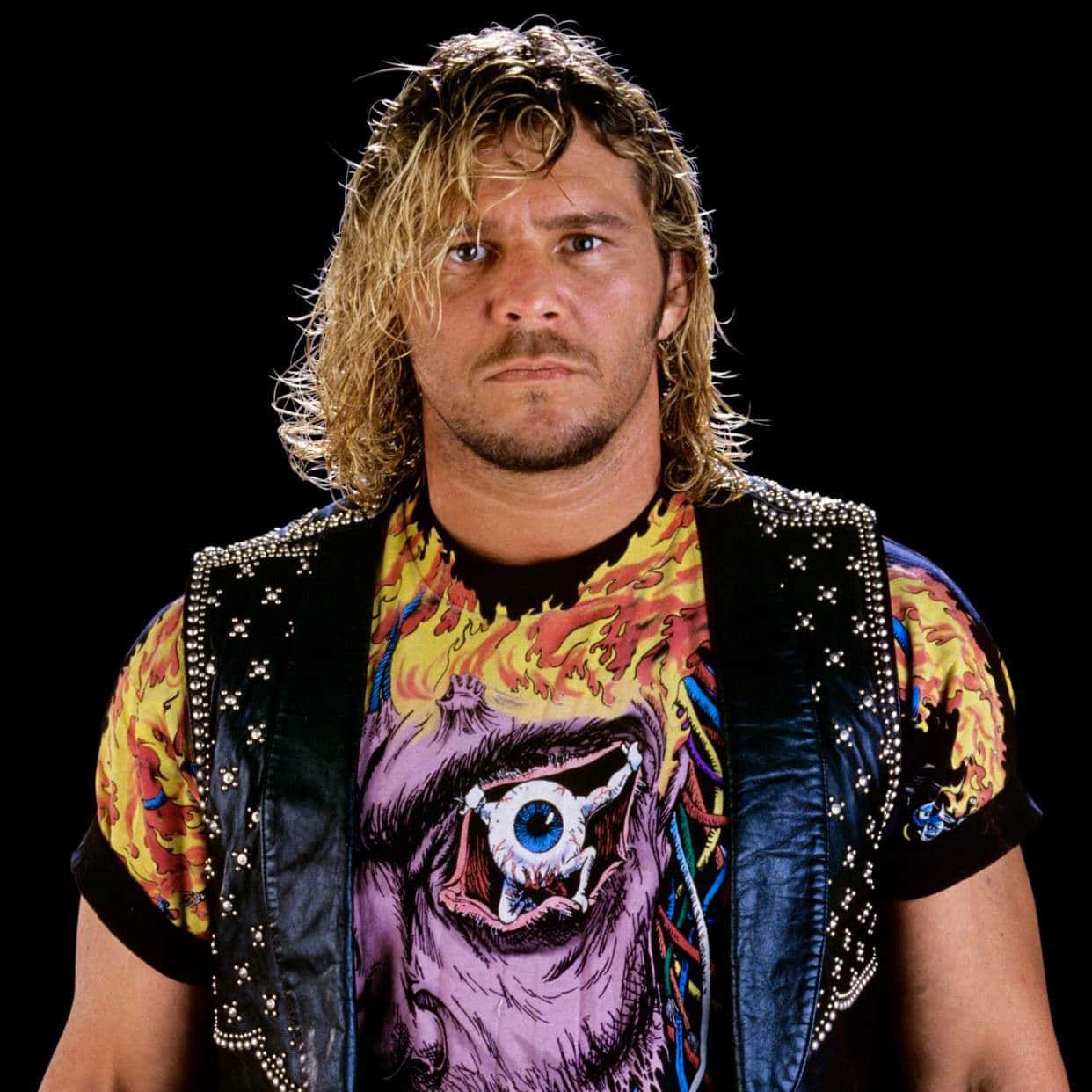WWE 桌布 壁紙 — 一位長金髮的摔角選手穿著釘珠背心，內搭印有怪獸眼睛圖案的圖案T恤，眼神銳利地凝視。