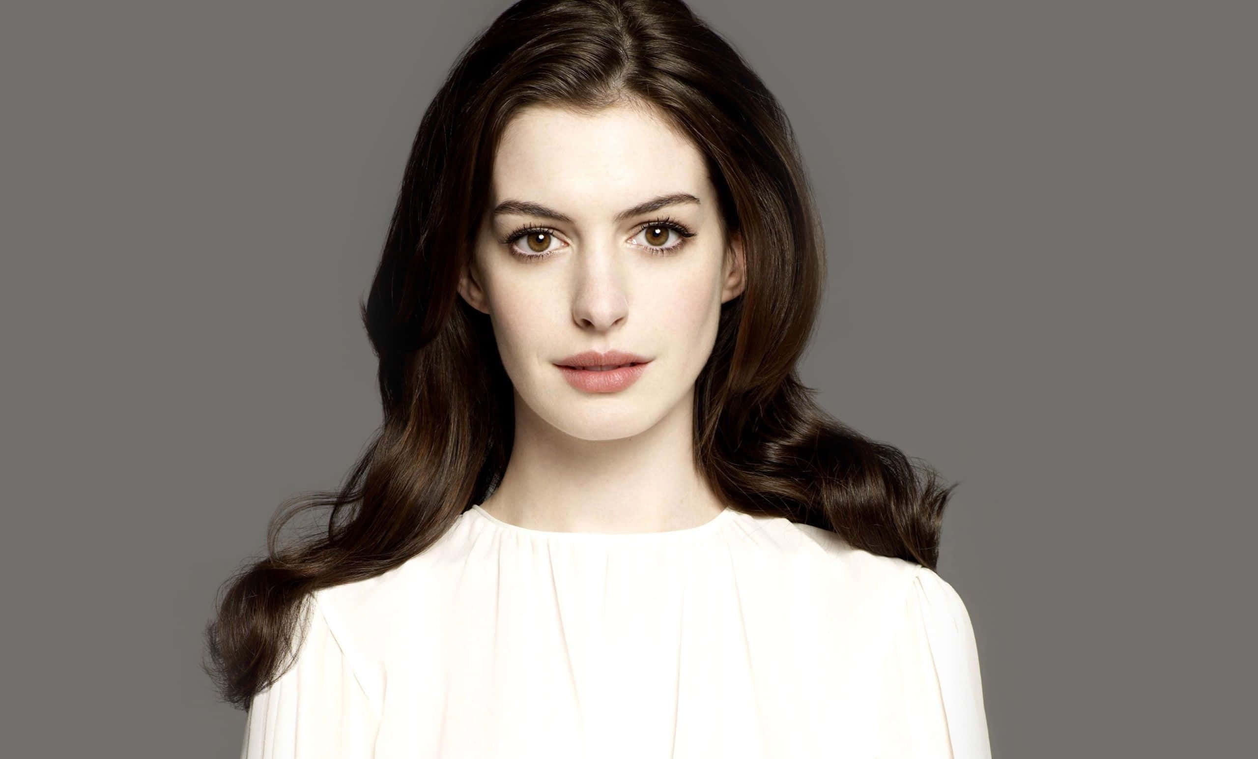 Anne Hathaway 桌布 壁紙 — 一位長長棕色頭髮、穿著白色襯衫的女性，在純灰色背景前平靜地凝視。