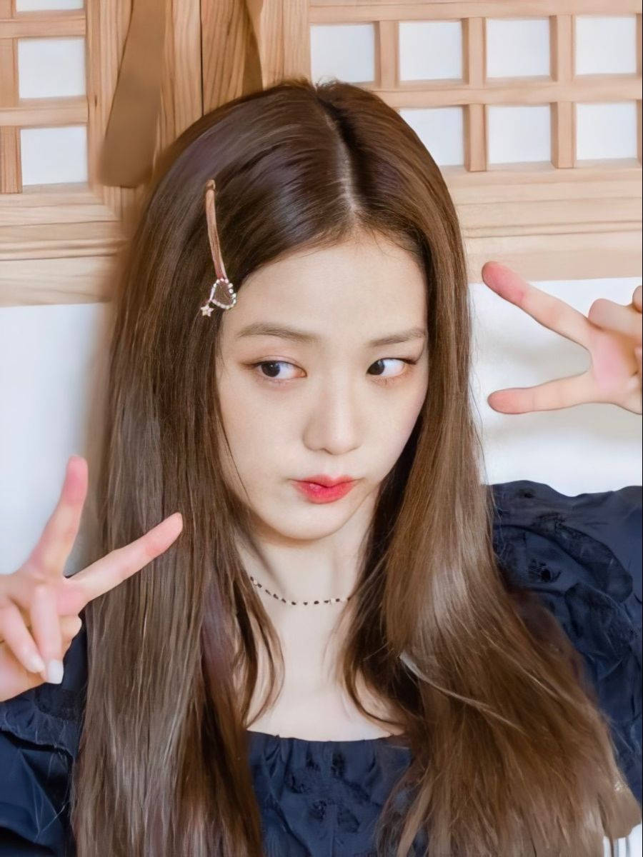 jisoo 可愛 桌布 壁紙 — 一位長長棕色頭髮和紅唇的年輕女性在木格柵背景前做出俏皮的和平手勢。