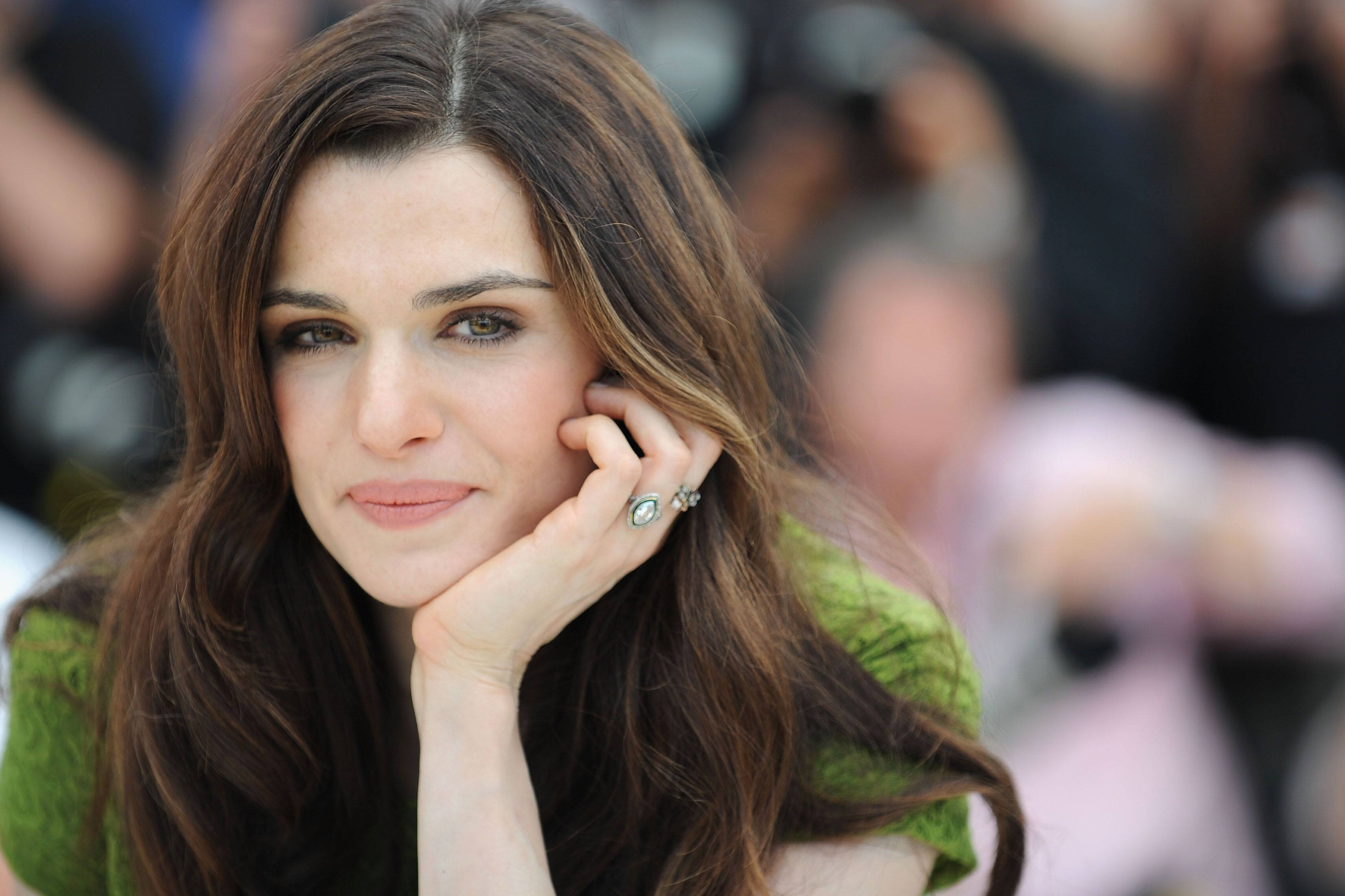 Rachel Weisz 桌布 壁紙 — 一位長長棕色頭髮的女性，用手托著下巴，穿著綠色上衣和戒指，看著相機。