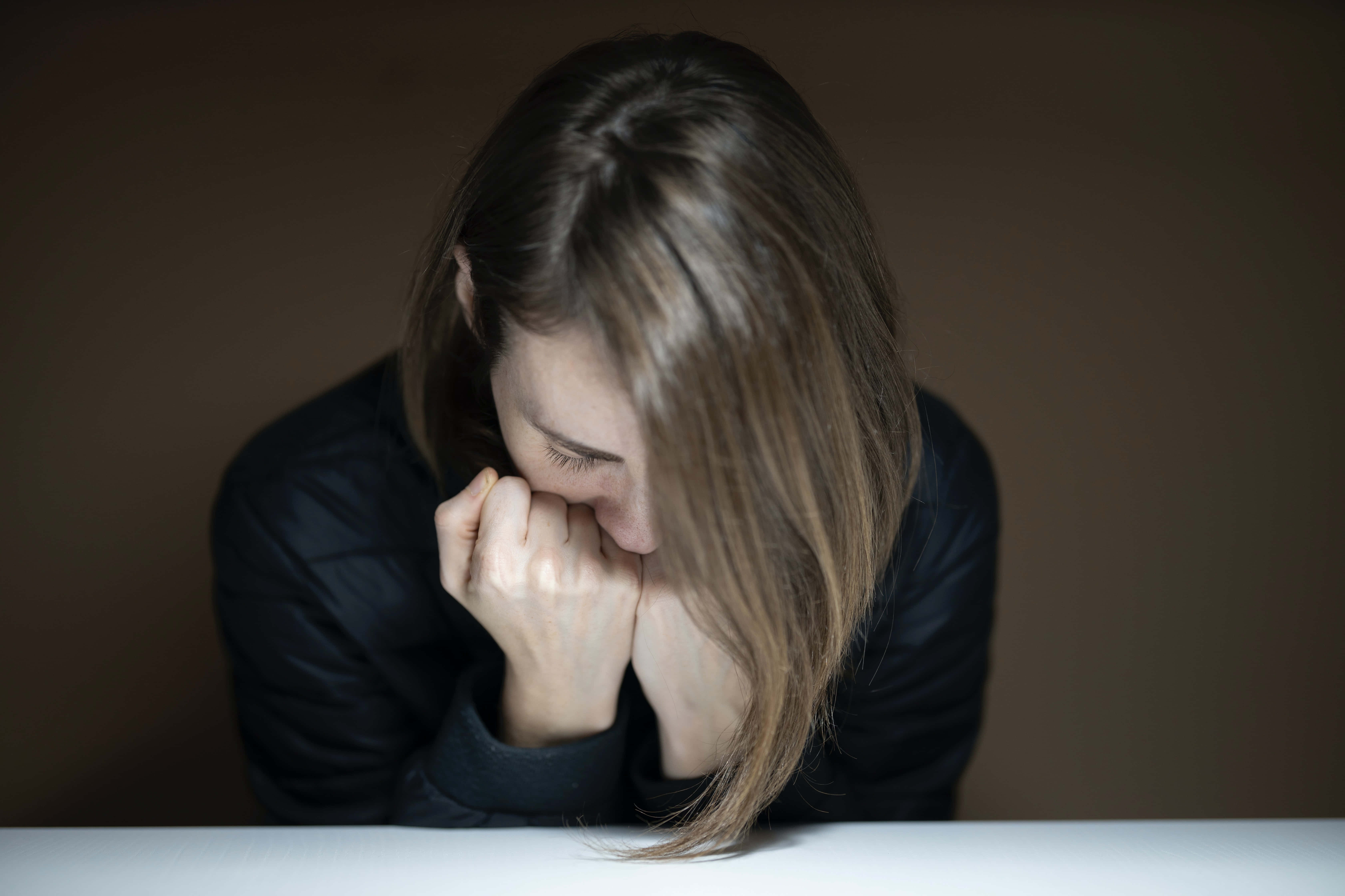 傷心 桌布 壁紙 — 一位長長棕色頭髮的女性在燈光昏暗的房間裡，將頭靠在白色桌子上緊握的雙手上。