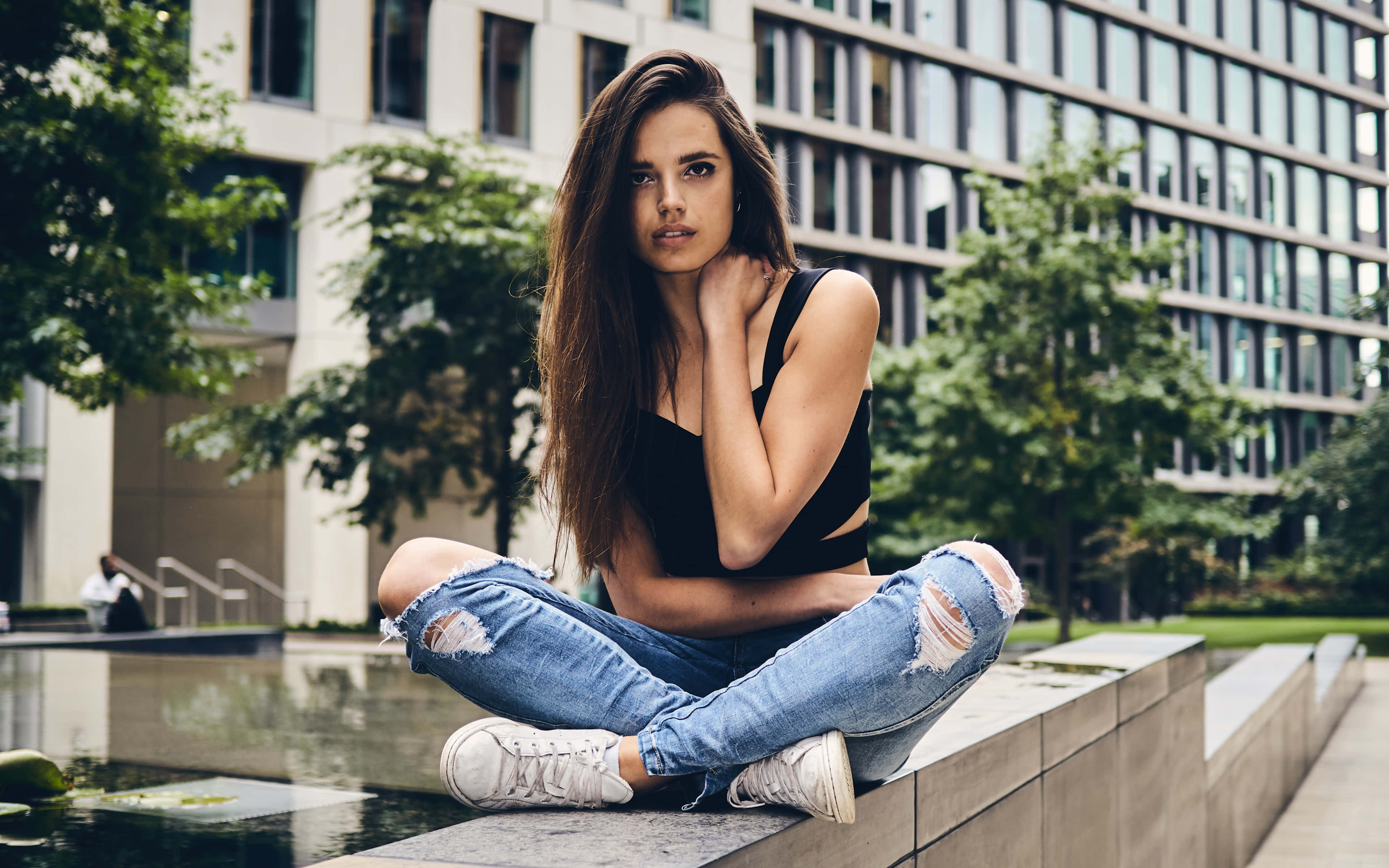 Sexy 桌布 壁紙 — 一位長長棕色頭髮的年輕女性，在現代城市廣場的水景旁，盤腿坐在混凝土邊緣上，穿著破洞牛仔褲和黑色上衣，直視鏡頭。