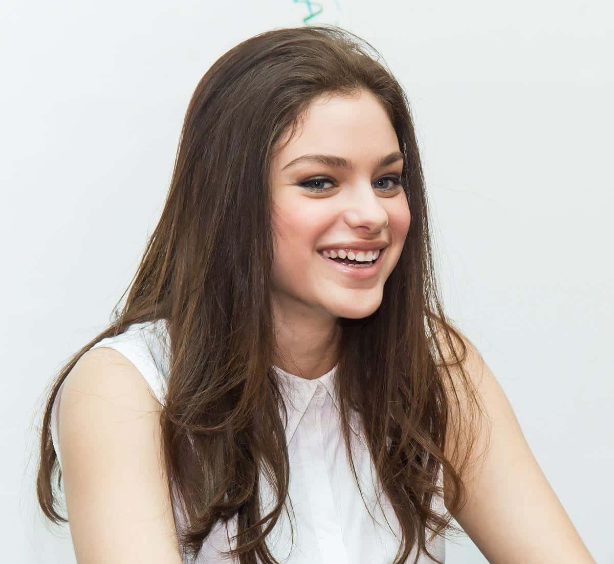 Odeya Rush 桌布 壁紙 — 一位長長棕色頭髮的年輕女性，穿著白色無袖上衣，在純白色背景前溫暖地微笑。