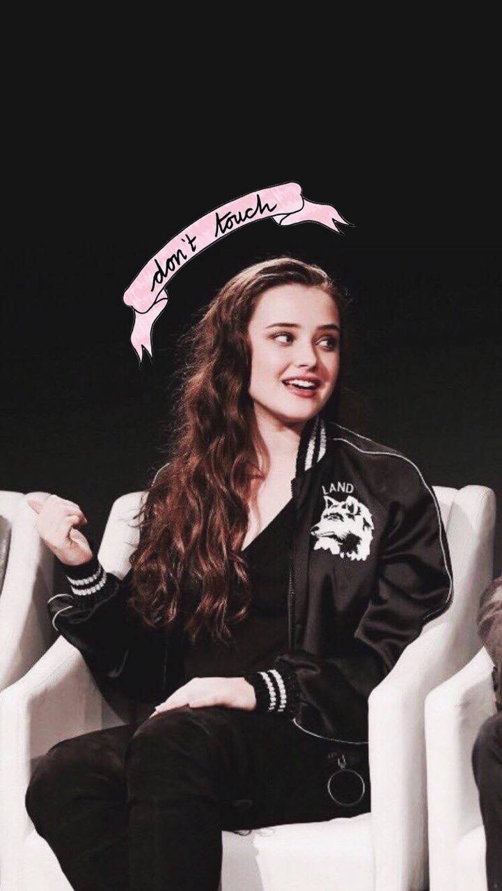 Katherine Langford 桌布 壁紙 — 一位長長棕色頭髮的年輕女性微笑著坐在白色椅子上，身穿帶有狼徽章的黑色飛行員夾克，頭頂上的粉紅色橫幅寫著「請勿觸摸」。