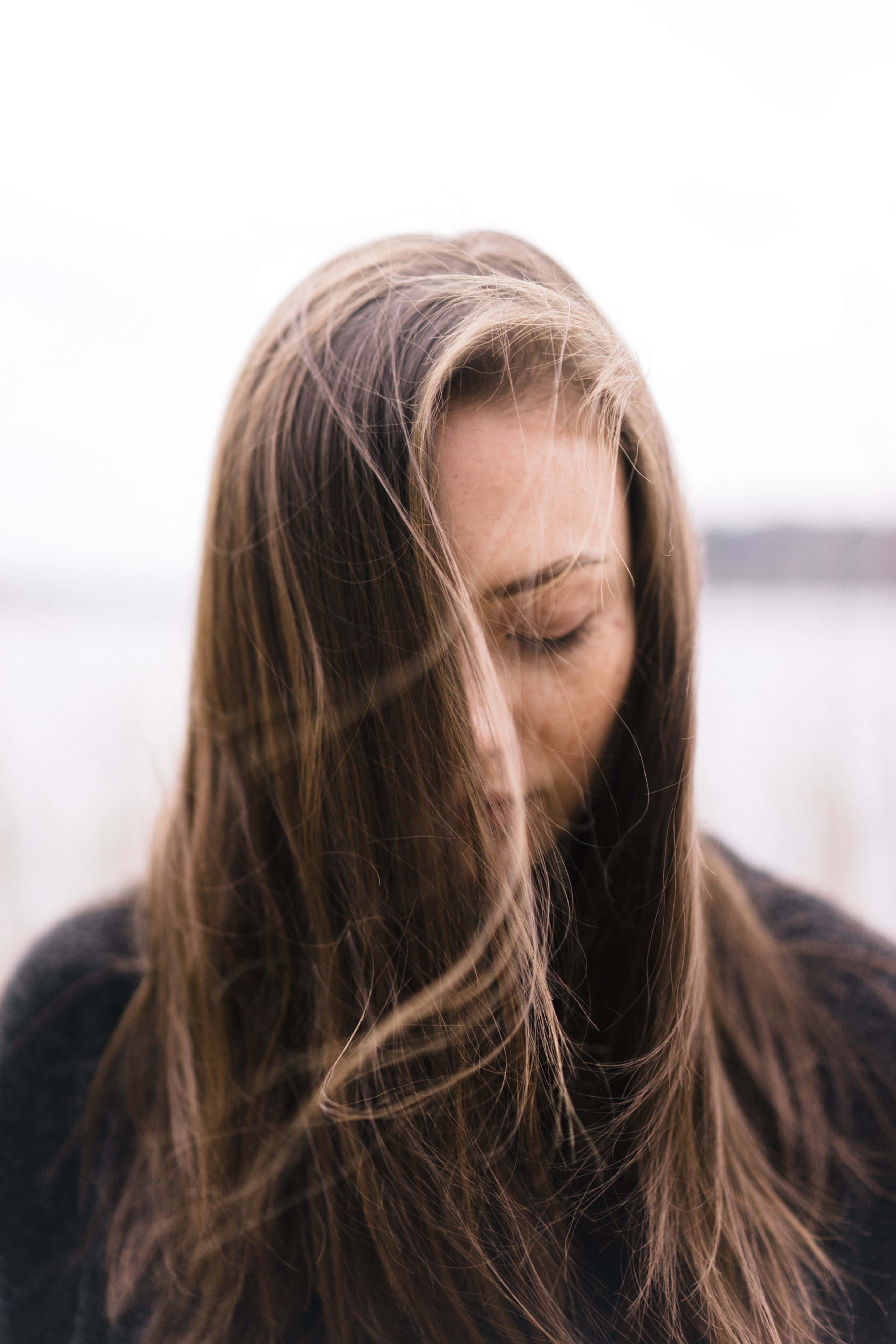 女孩 桌布 壁紙 — 一位長長棕色頭髮隨風輕柔飄揚的女性，在柔和模糊的背景前閉上眼睛，平靜地沉思。