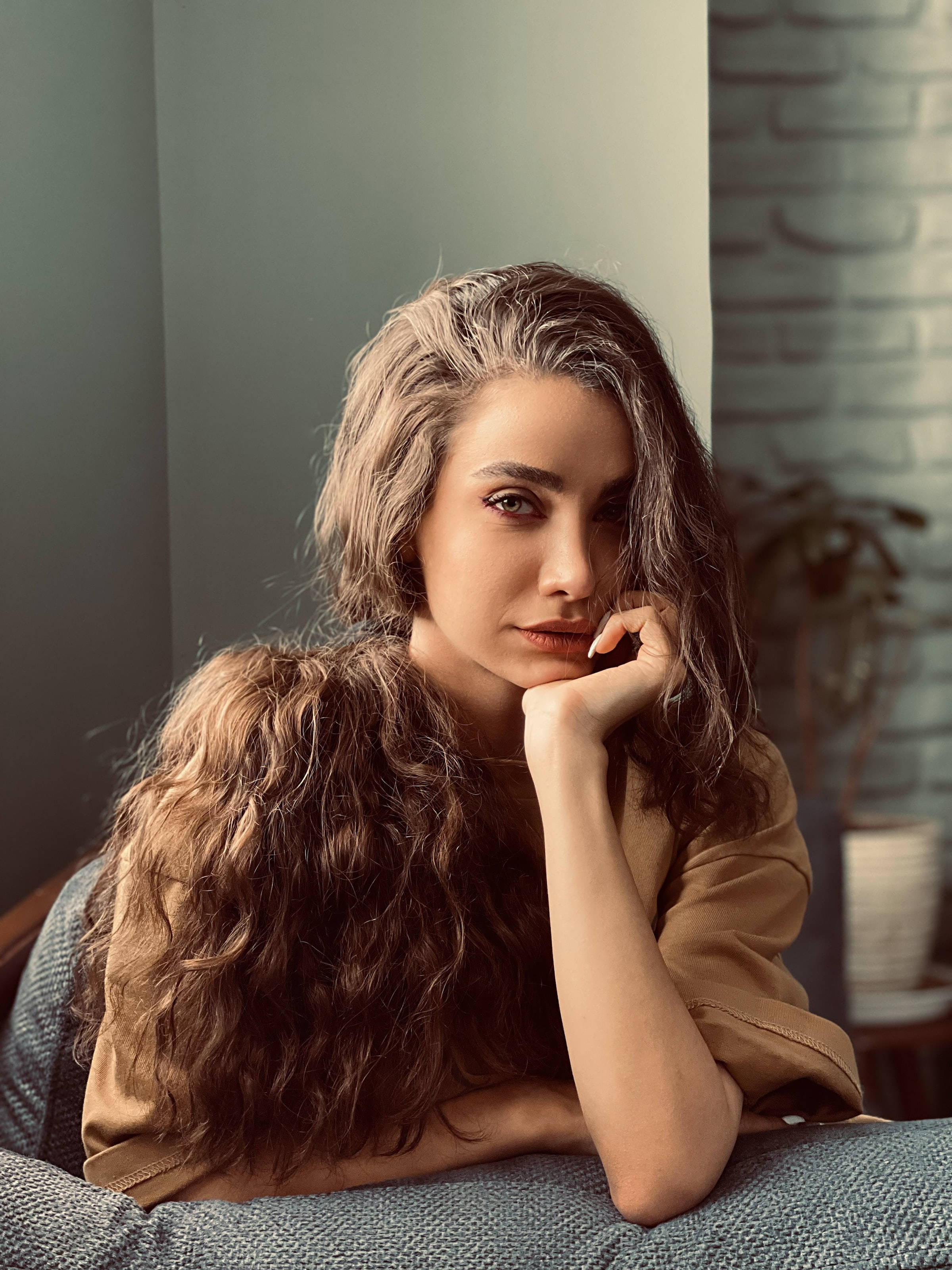 female 桌布 壁紙 — 一位長長波浪棕色頭髮的女性，在柔和光線的現代室內環境中，坐在有紋理的沙發上，用手托著下巴。