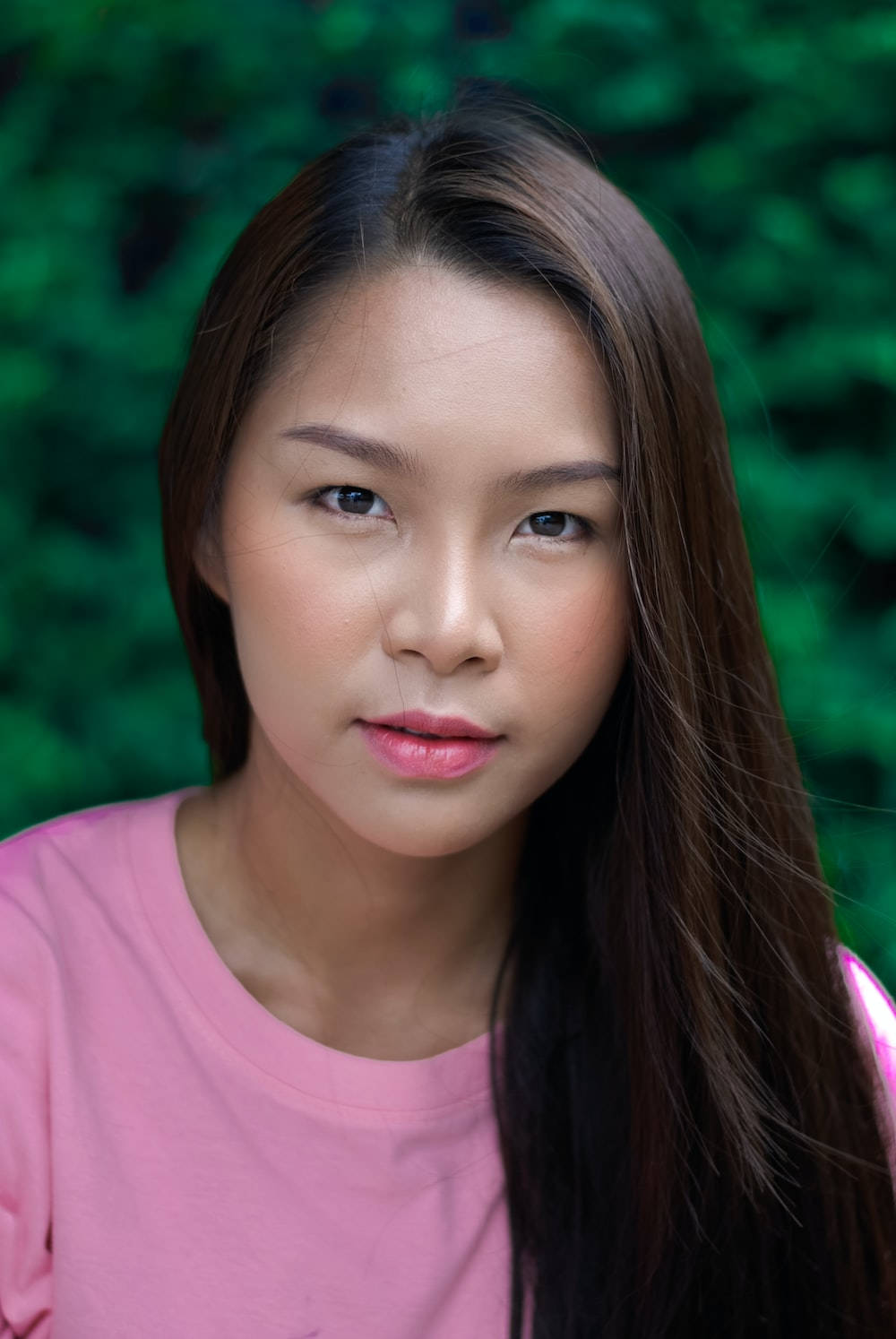Thailand Woman 桌布 壁紙 — 一位長長深色頭髮、穿著粉紅色上衣的年輕女性，在茂密的綠色樹葉背景下溫柔地凝視著。