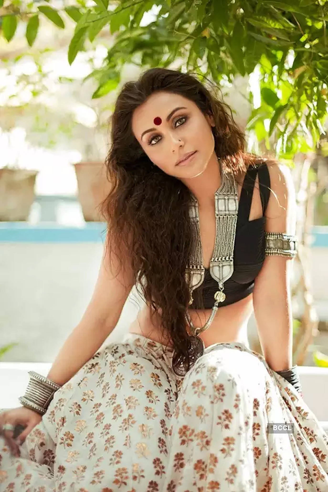 Rani Mukerji 桌布 壁紙 — 一位長長深色頭髮、額頭有紅色吉祥痣的女性，優雅地坐在綠樹成蔭下，佩戴華麗的銀飾和花卉裙。