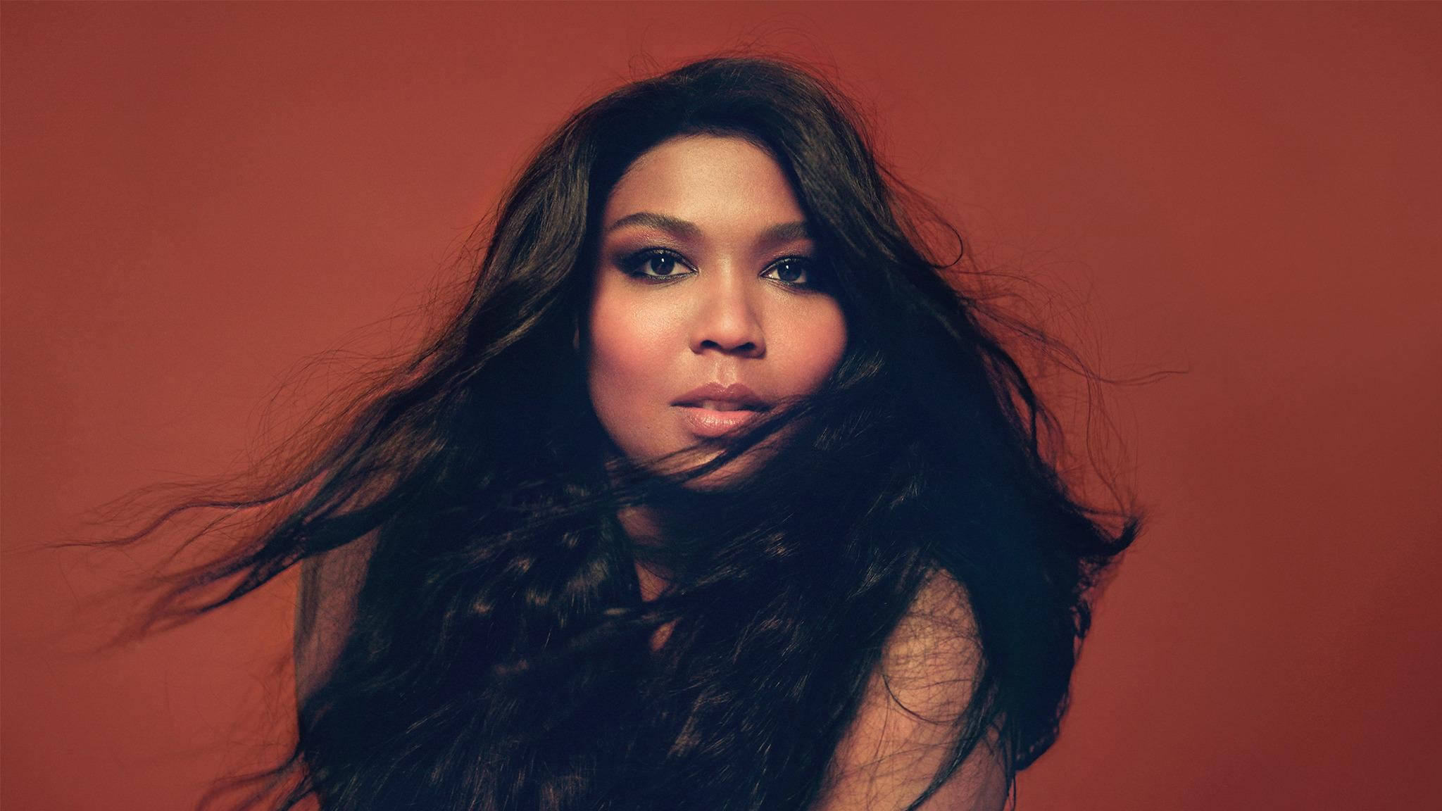 Lizzo 桌布 壁紙 — 一位長長深色頭髮的女性，頭髮在溫暖的陶土色背景前吹過她的臉龐。