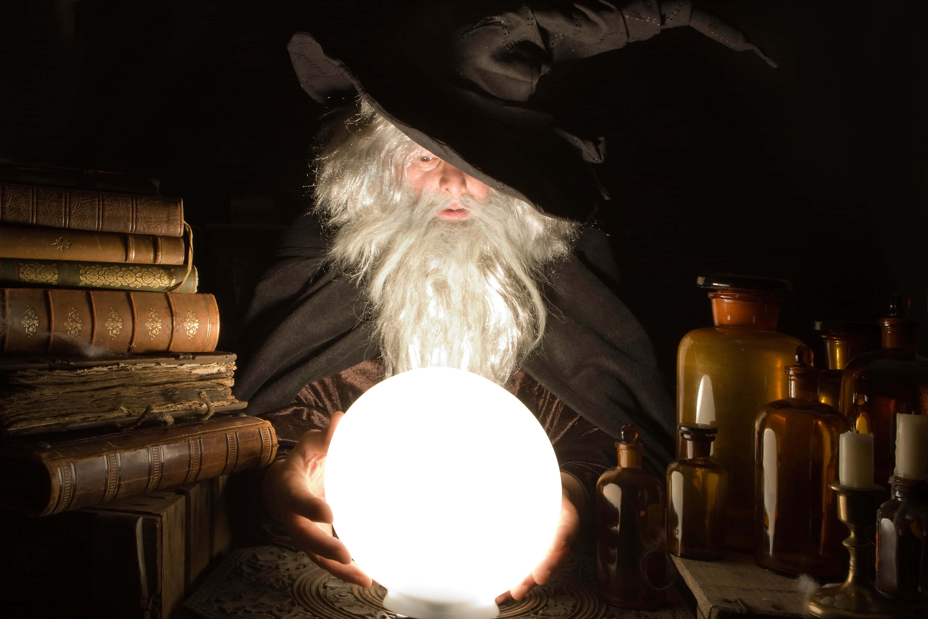 Fortune Teller 桌布 壁紙 — 一位長長白鬍子、戴黑帽的巫師，在一個光線昏暗、佈滿古籍和琥珀色玻璃瓶的書房中，手持一顆發光的水晶球。