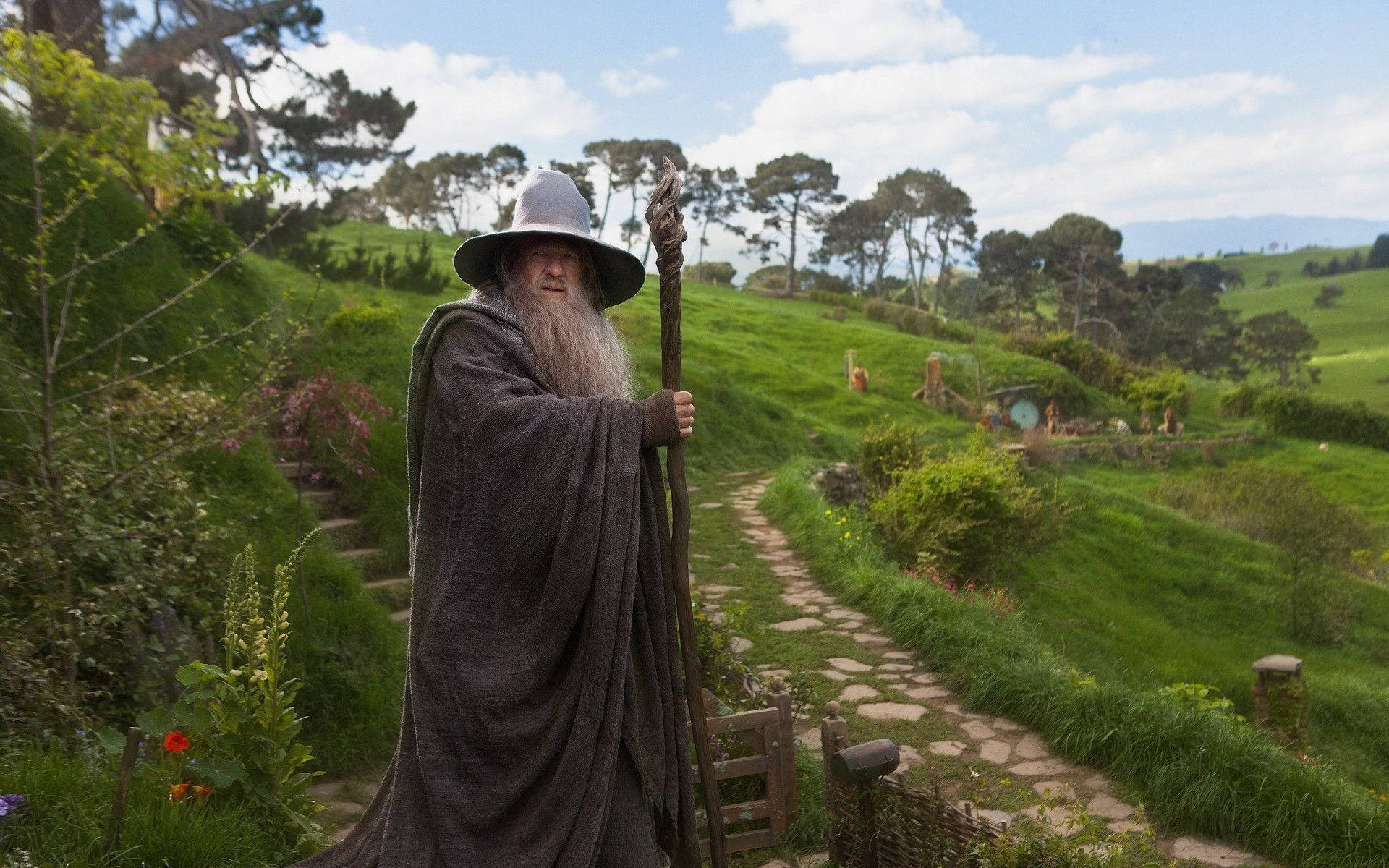 the hobbit 桌布 壁紙 — 一位長長白鬍鬚、身穿灰色長袍的年老巫師，站在翠綠哈比村莊的石徑上，頭頂是明亮的藍天。