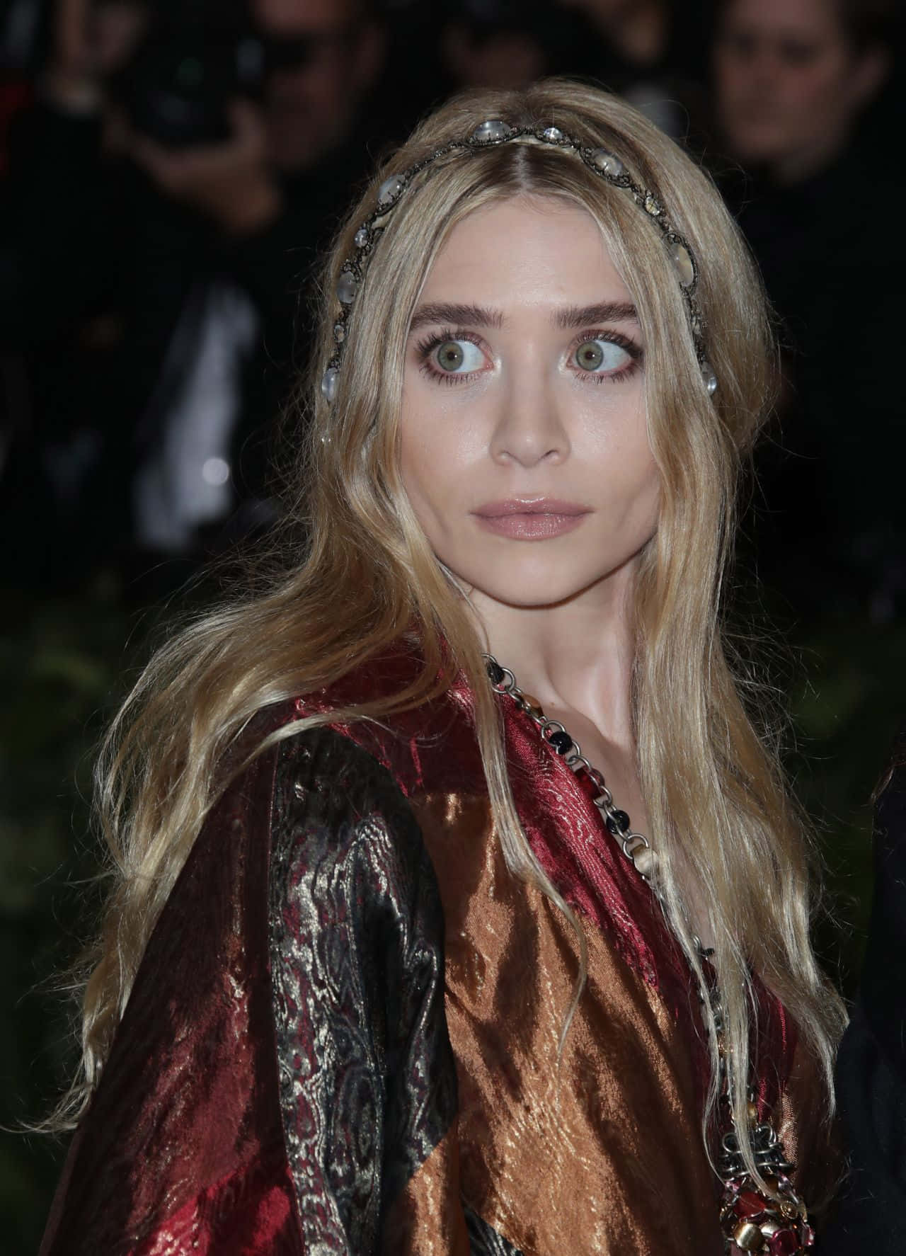 Mary Kate Olsen 桌布 壁紙 — 一位長長金髮、戴著珠寶頭帶的女性，身穿閃亮的紅金色服裝，在擁擠的活動中凝視前方。