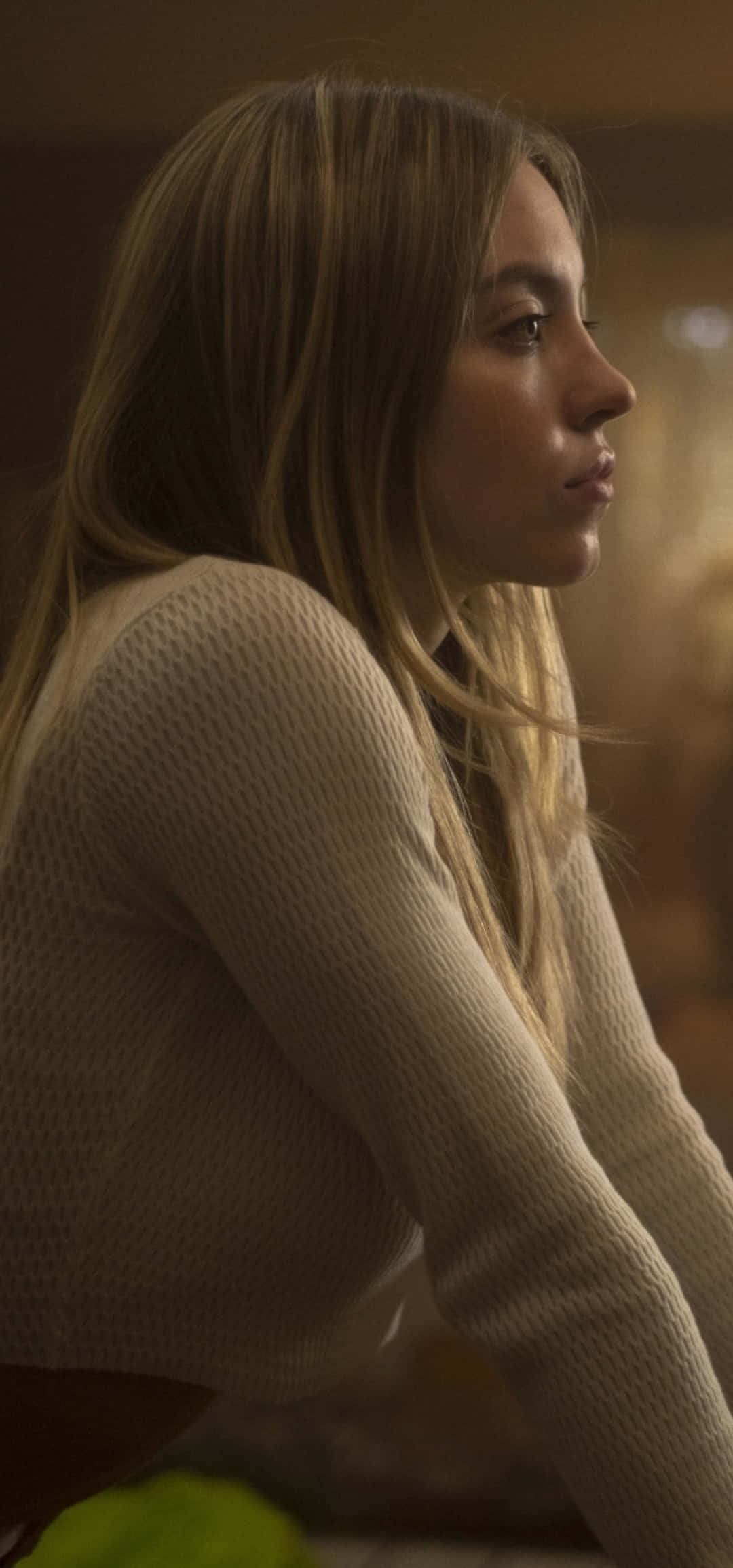 Euphoria Hbo iPhone 桌布 壁紙 — 一位長長金髮的年輕女性坐在溫暖的室內光線中，穿著一件有質感的奶油色毛衣。