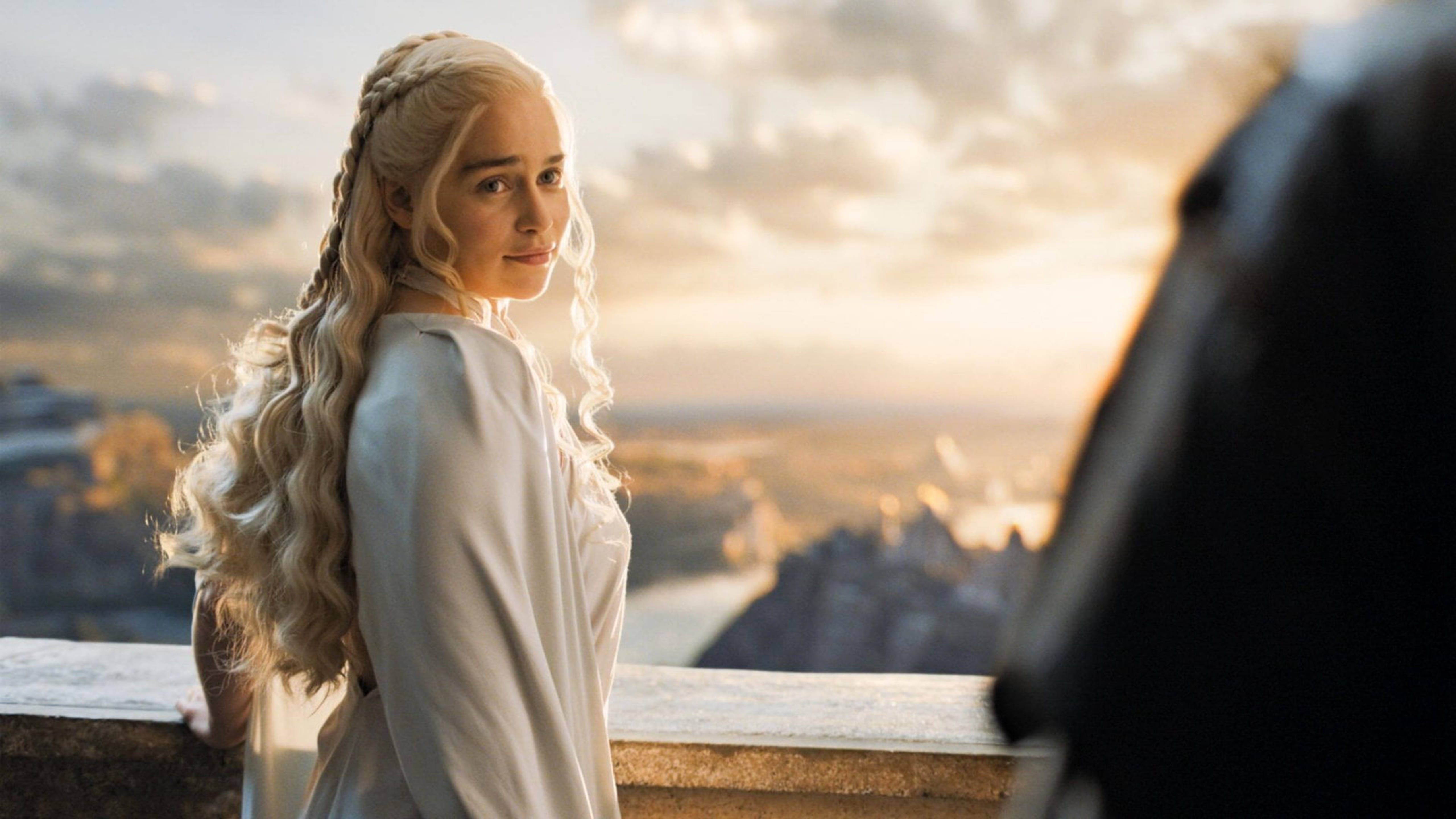 daenerys 桌布 壁紙 — 一位長長金髮辮子、身穿白色長袍的女子，站在石造陽台上，俯瞰金色夕陽下蔓延的城市。
