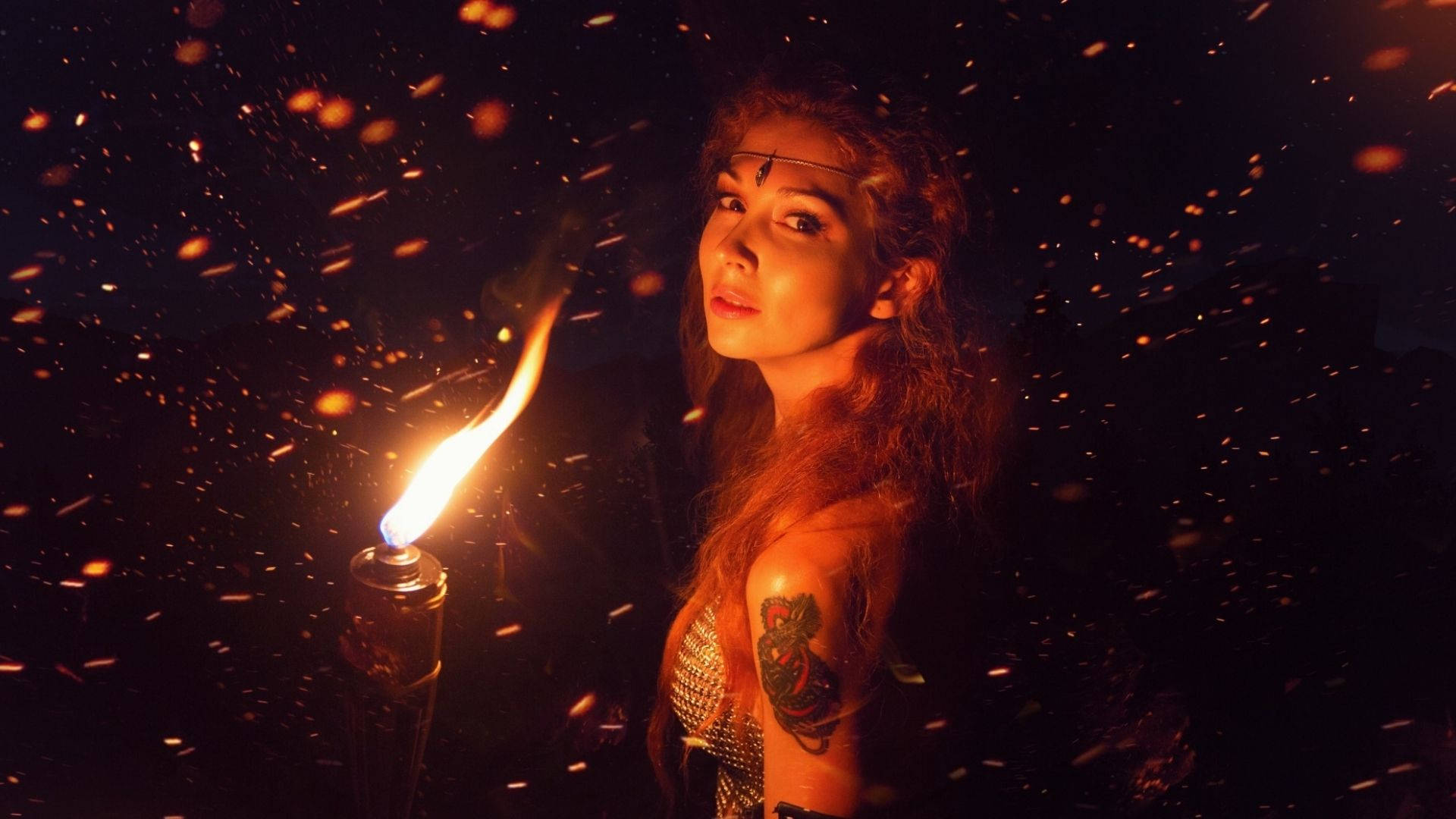 Fire Girl 桌布 壁紙 — 一位長髮且有刺青的女性在黑暗中仰望，火花四濺，手持燃燒的火炬。