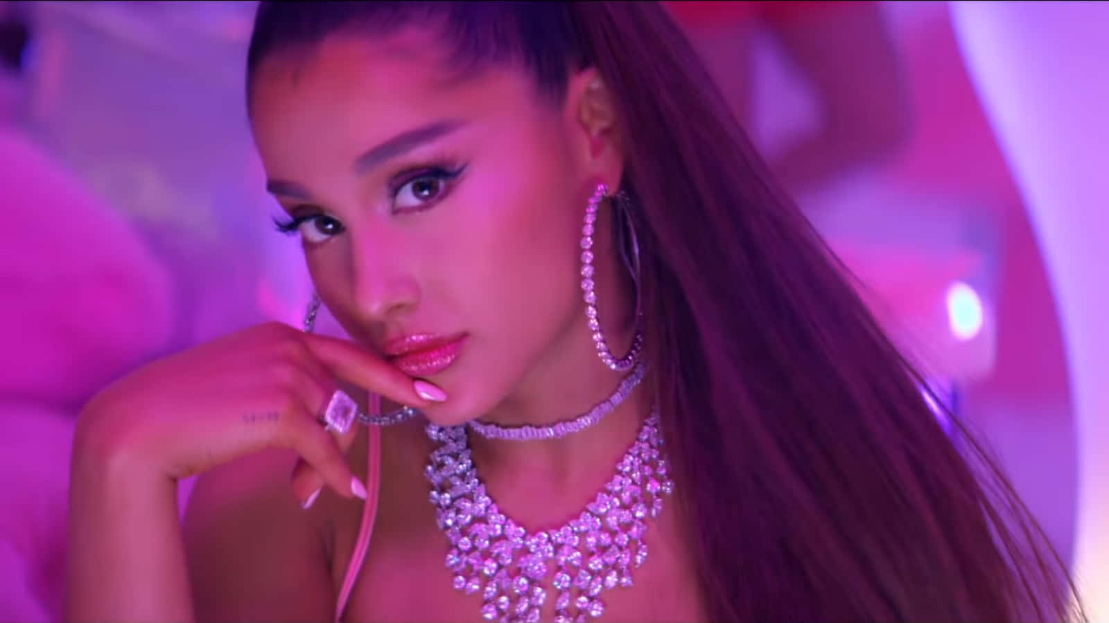 Ariana Grande 7 Rings 桌布 壁紙 — 一位長髮女子佩戴鑽石首飾，在充滿活力的粉紅燈光下自信地擺姿勢。