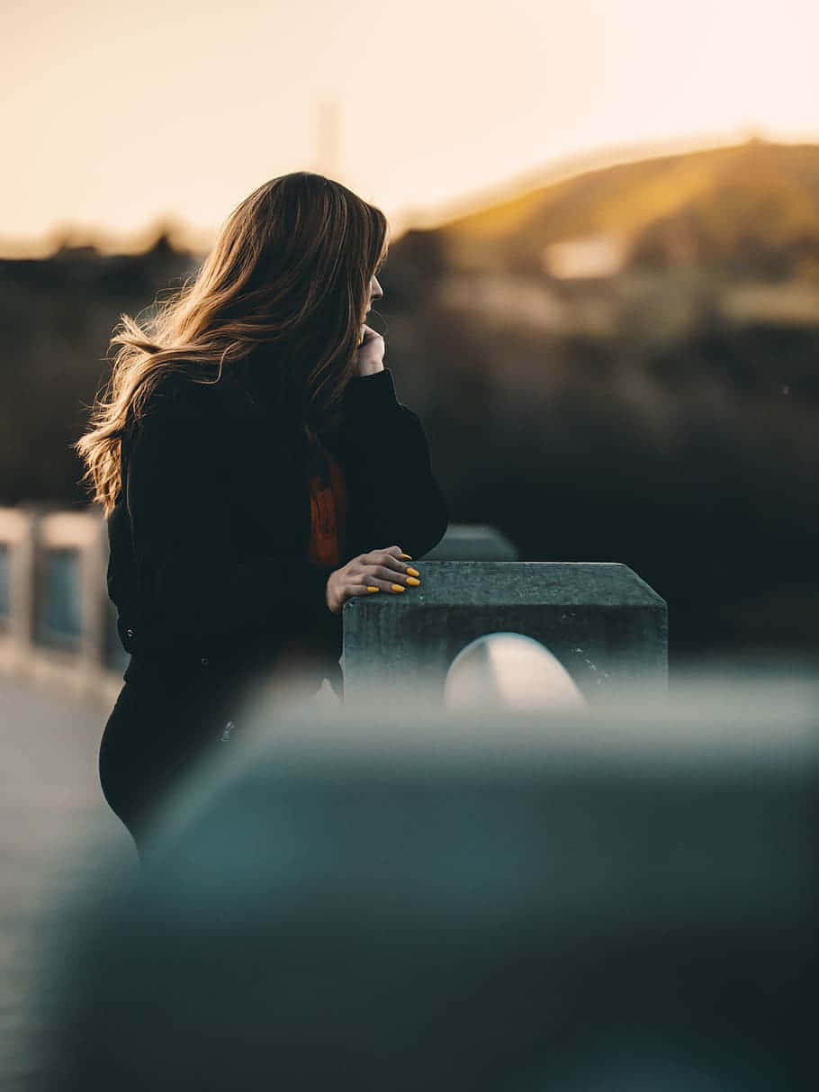 Woman 桌布 壁紙 — 一位長髮女子在日落時倚靠在石欄杆上，她的剪影被溫暖的金色光芒框住，背景是模糊的山丘。