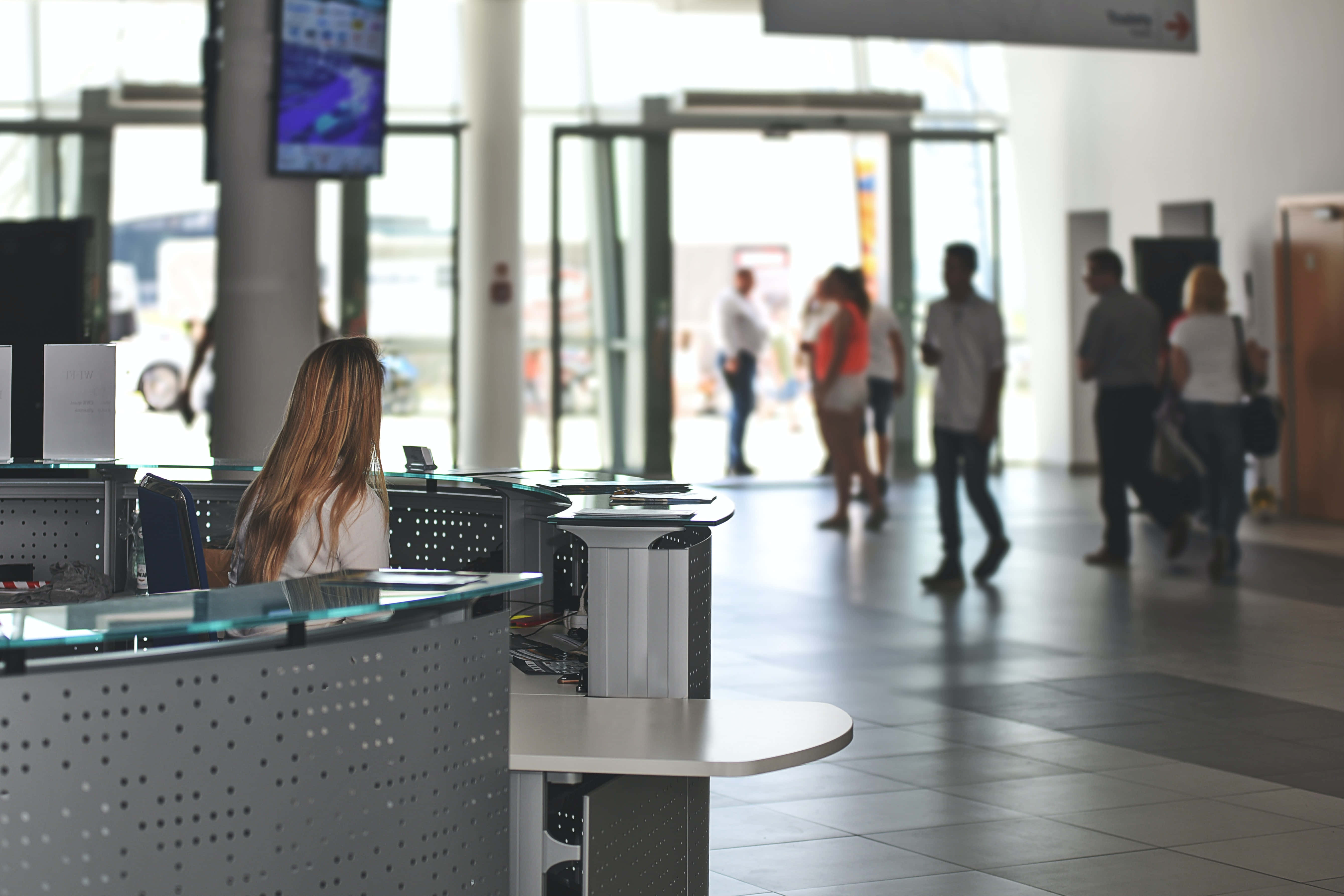Airport 桌布 壁紙 — 一位長髮女性坐在明亮寬敞的機場航廈內現代化的玻璃和金屬服務櫃檯，模糊的旅客走過。