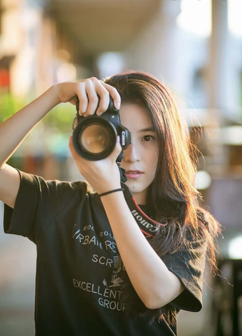 Holding Camera 桌布 壁紙 — 一位長髮年輕女性在陽光明媚的城市環境中，將Canon DSLR相機舉到眼前。