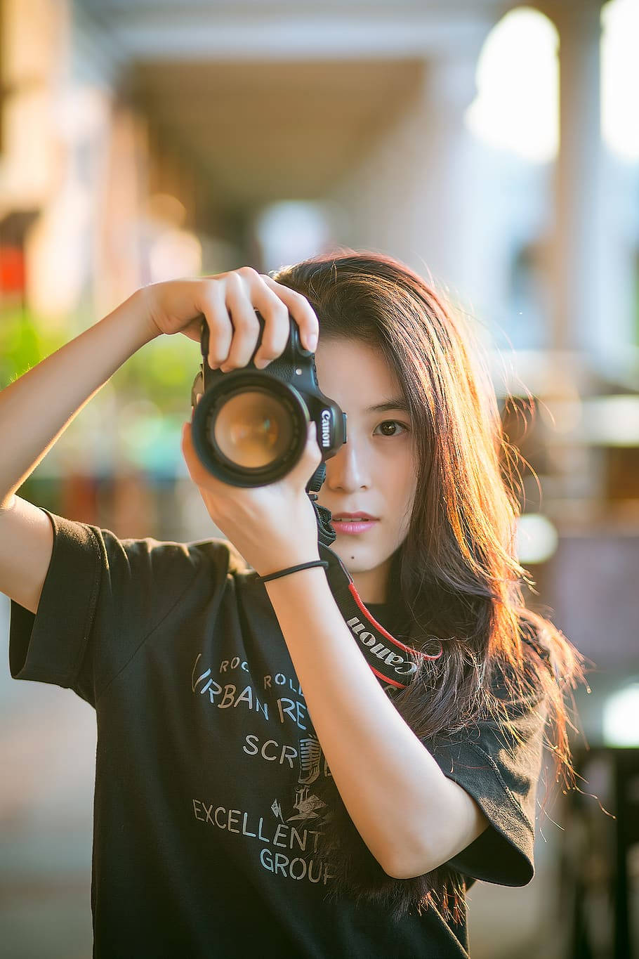 Woman 桌布 壁紙 — 一位長髮年輕女性在陽光普照的城市小巷中，將Canon DSLR相機舉到眼前。