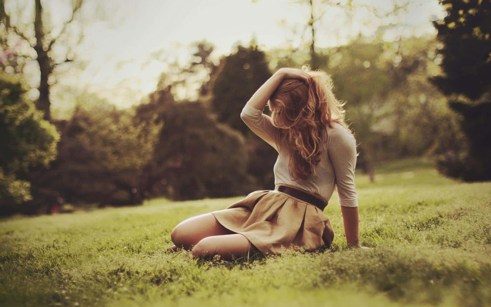 Sad Romance 桌布 壁紙 — 一位長髮年輕女性坐在陽光普照的公園草地上，手放在頭上，被溫暖金色光線中的樹木環繞。