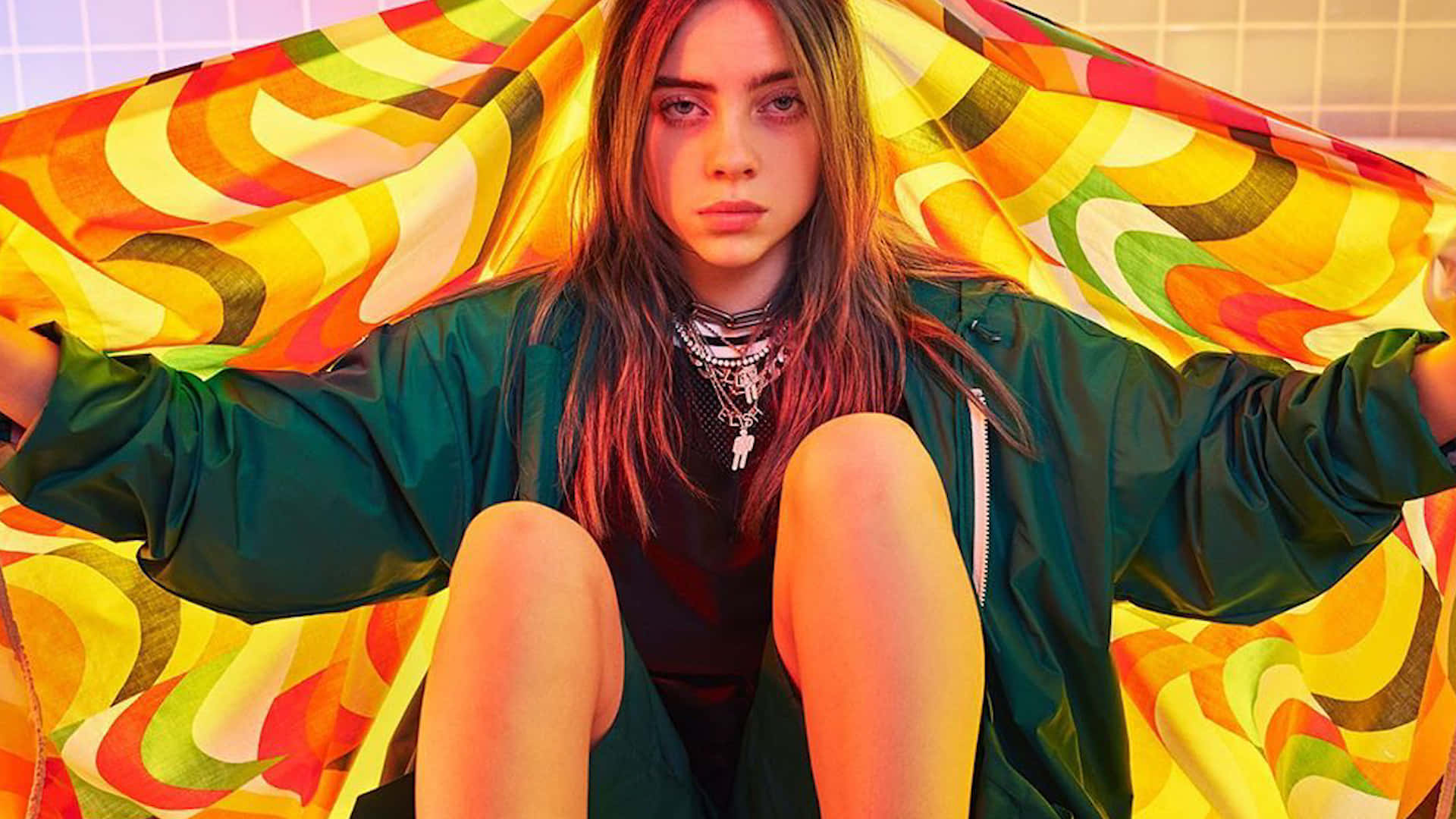 Billie Eilish Laptop 桌布 壁紙 — 一位長髮年輕女性自信地坐著，手持色彩鮮豔、漩渦圖案的布料，靠著瓷磚牆。