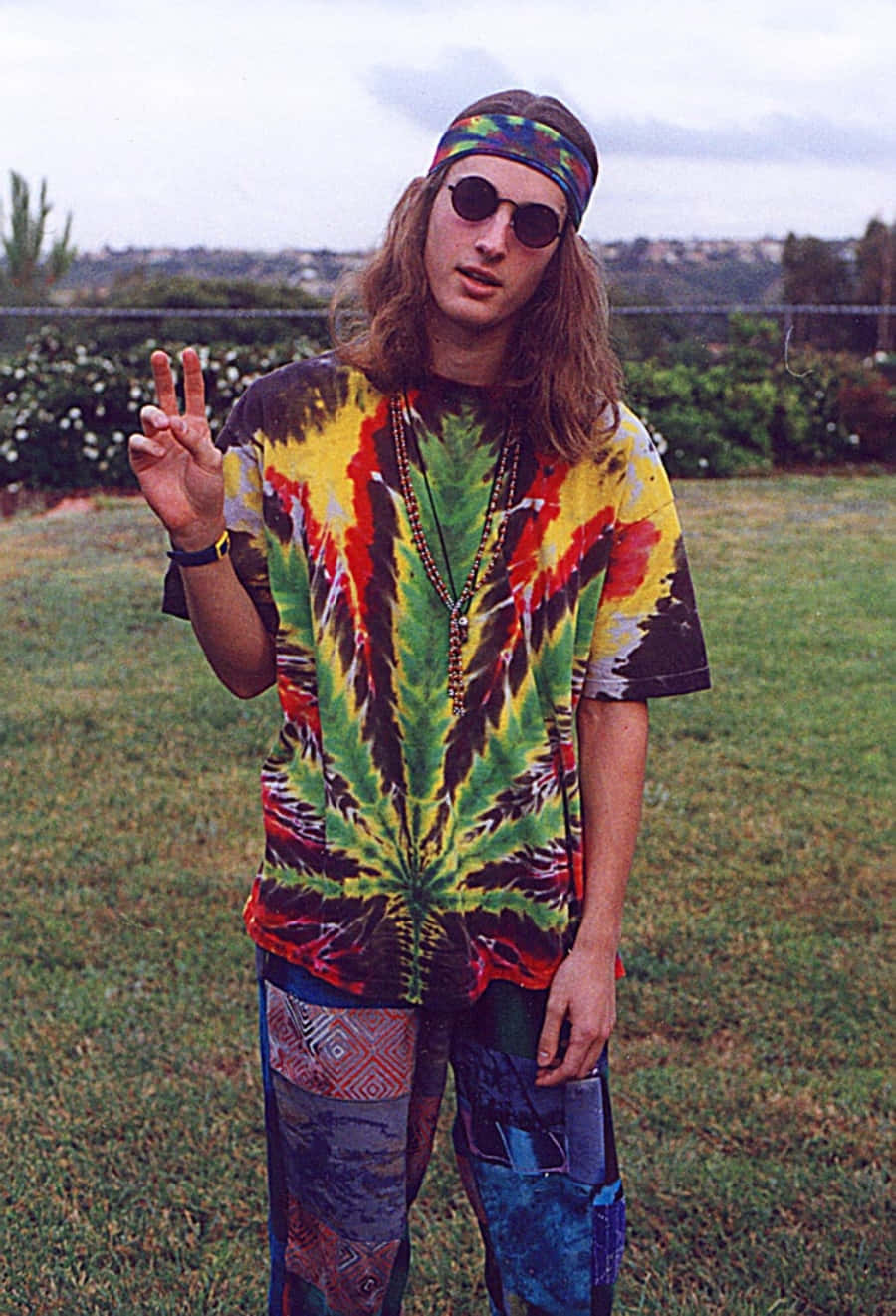 嬉皮 桌布 壁紙 — 一位長髮戴太陽眼鏡的年輕男子穿著鮮豔的紮染襯衫和圖案褲子，在戶外比出和平手勢。