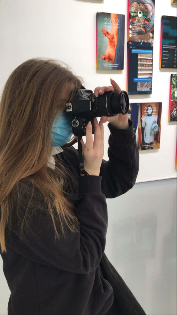 Holding Camera 桌布 壁紙 — 一位長髮戴著口罩的人，站在展示色彩繽紛手機桌布海報的牆前，手持DSLR相機。