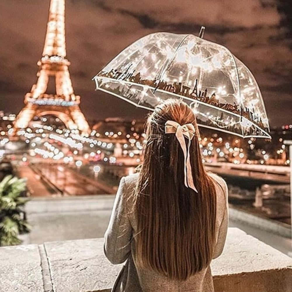 Eiffel Tower 桌布 壁紙 — 一位長髮戴蝴蝶結的女性，手持一把裝飾著巴黎天際線燈光的透明雨傘，凝視著夜晚燈火通明的艾菲爾鐵塔。