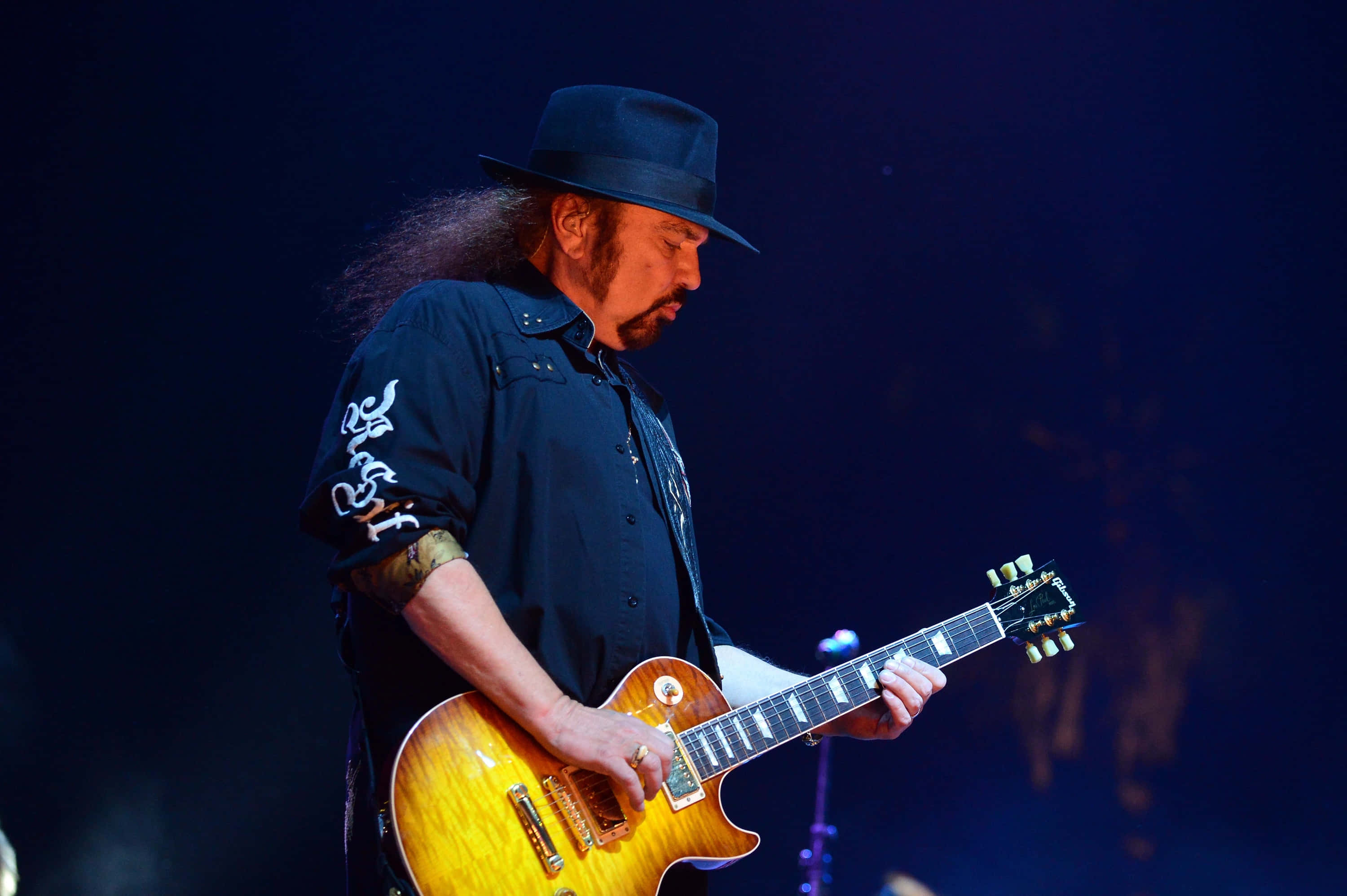Gary Rossington 桌布 壁紙 — 一位長髮戴黑帽的音樂家在舞台燈光下彈奏一把旭日紋電吉他。