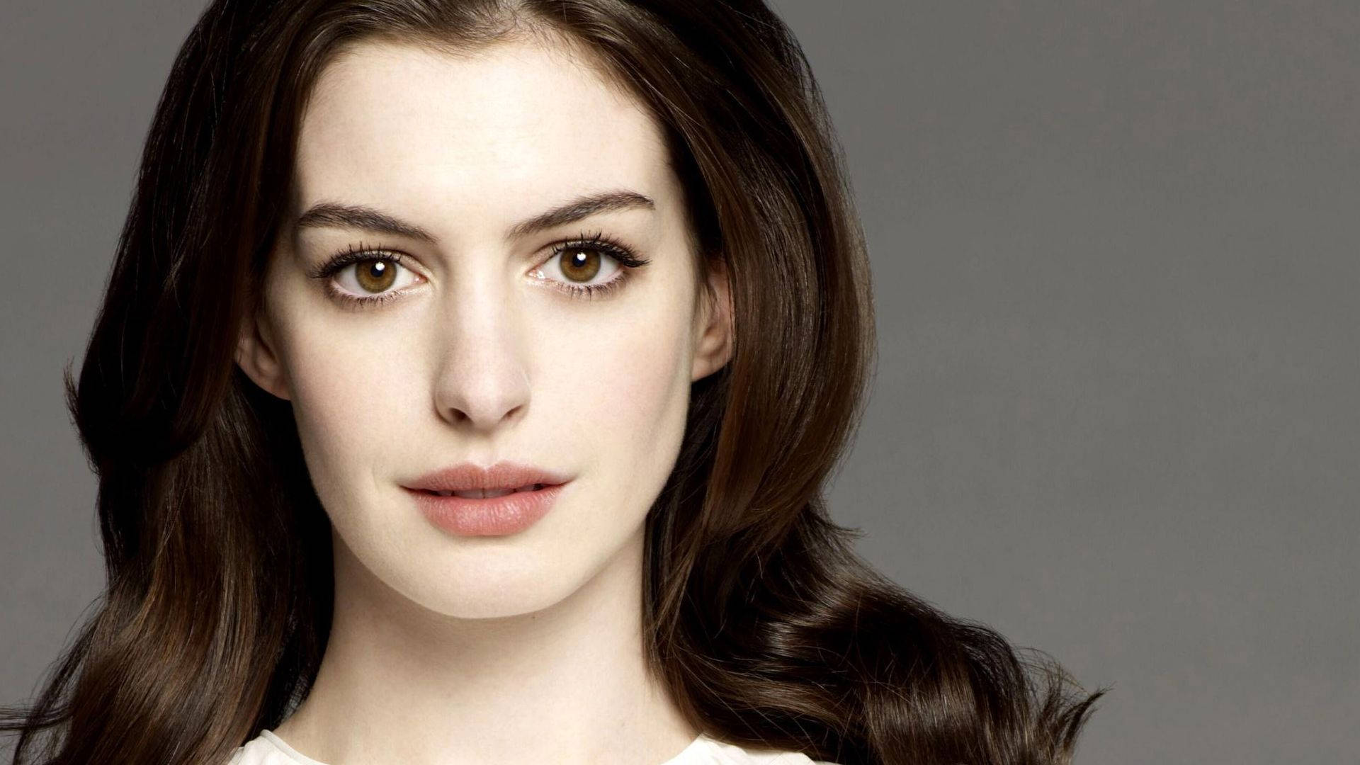 Anne Hathaway 桌布 壁紙 — 一位長髮棕眼、眼神溫暖的女性特寫肖像，直視鏡頭，背景為中性灰色。