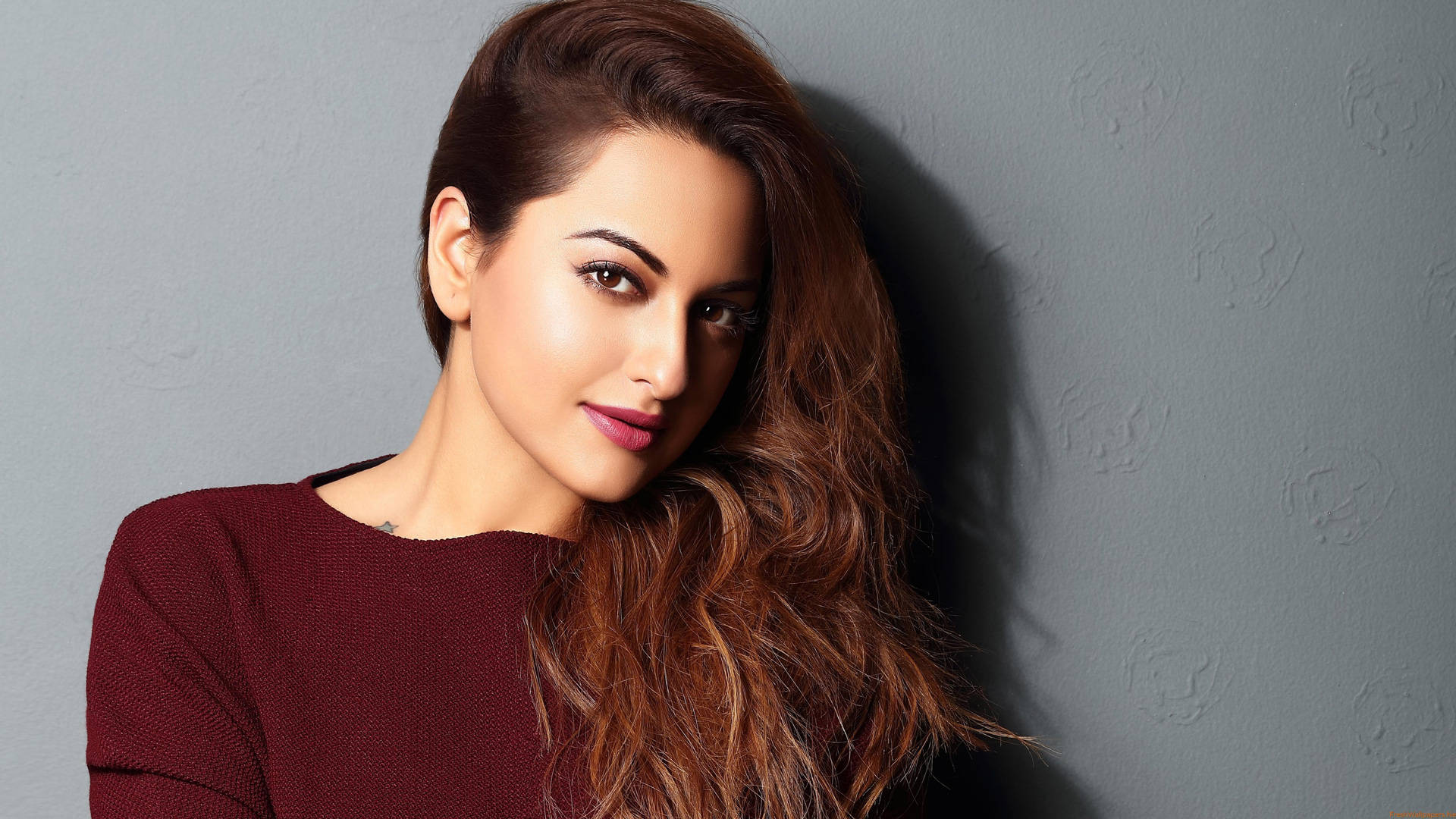 Sonakshi Sinha 桌布 壁紙 — 一位長髮棕眼、身穿紅色毛衣的女性，在柔和燈光下靠著紋理灰色牆壁。