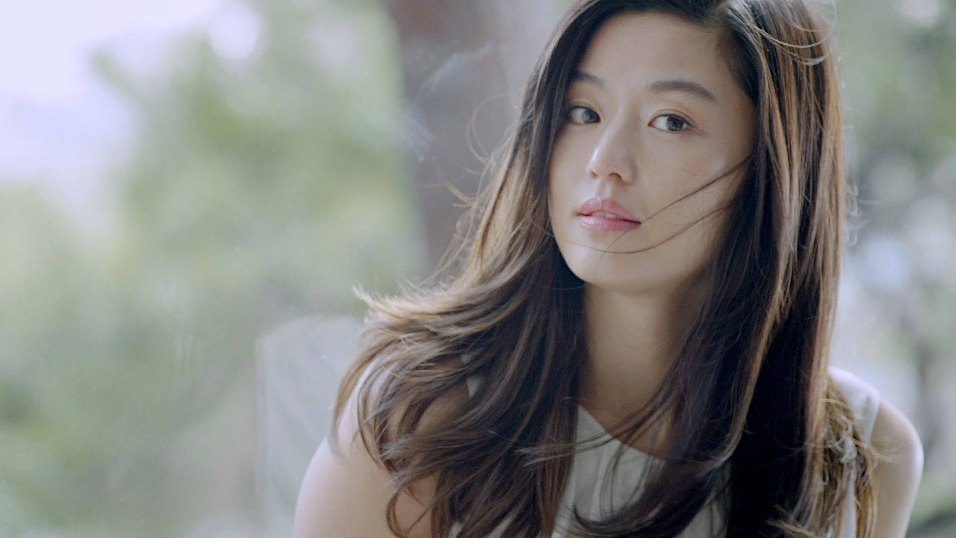 Jun Ji Hyun 桌布 壁紙 — 一位長髮棕眼的年輕女子，在模糊的綠色背景下，微風吹起她的髮絲，她若有所思地望著窗外。