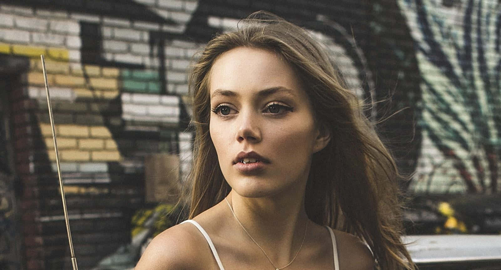 Grace Van Dien 桌布 壁紙 — 一位長髮棕眼的年輕女性在城市環境中，對著佈滿塗鴉的磚牆凝視。