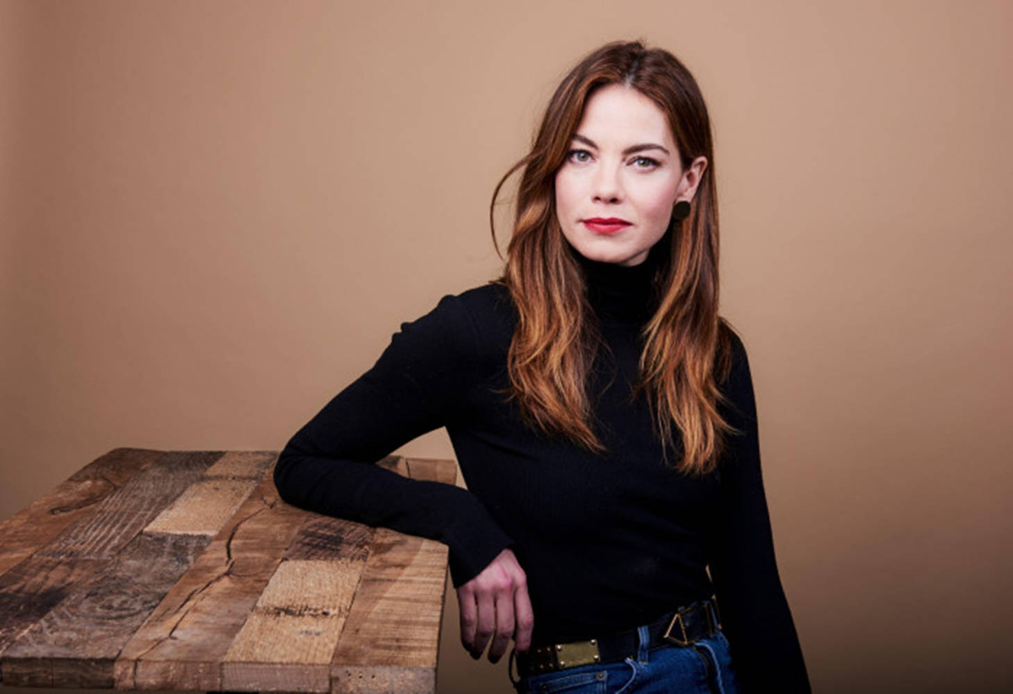 Michelle Monaghan 桌布 壁紙 — 一位長髮棕色的女士靠在一張質樸的木桌上，背景是溫暖的米色攝影棚。