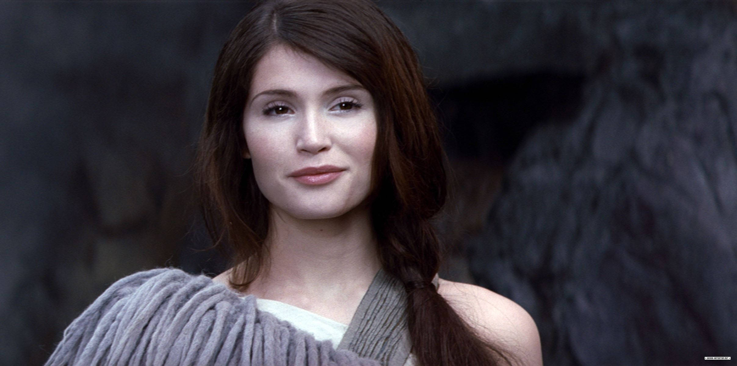Gemma Arterton 桌布 壁紙 — 一位長髮棕色的女性，披著灰色針織披肩，在黑暗的岩石背景下溫柔地凝視。