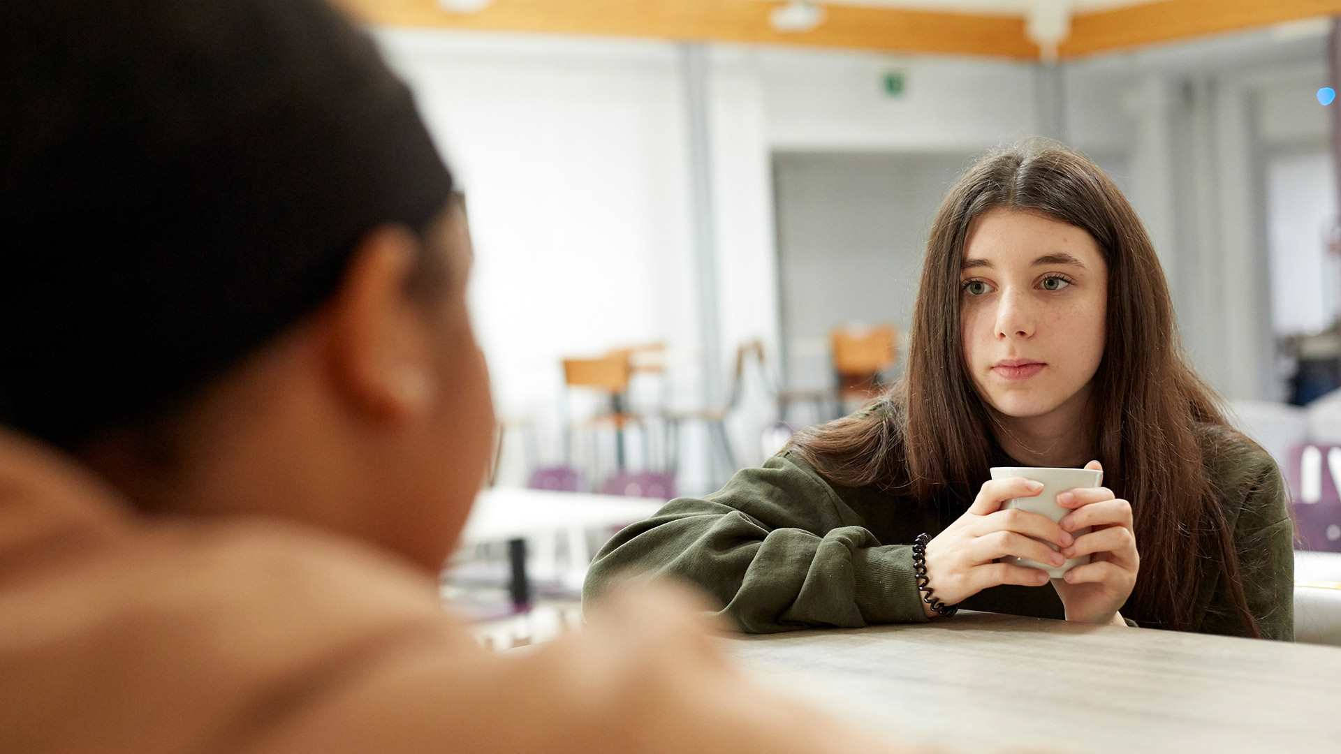 Anxiety 桌布 壁紙 — 一位長髮棕色的年輕女子坐在桌子旁，拿著一個白色杯子，專注地看著對面的另一個人，在明亮的室內環境中。
