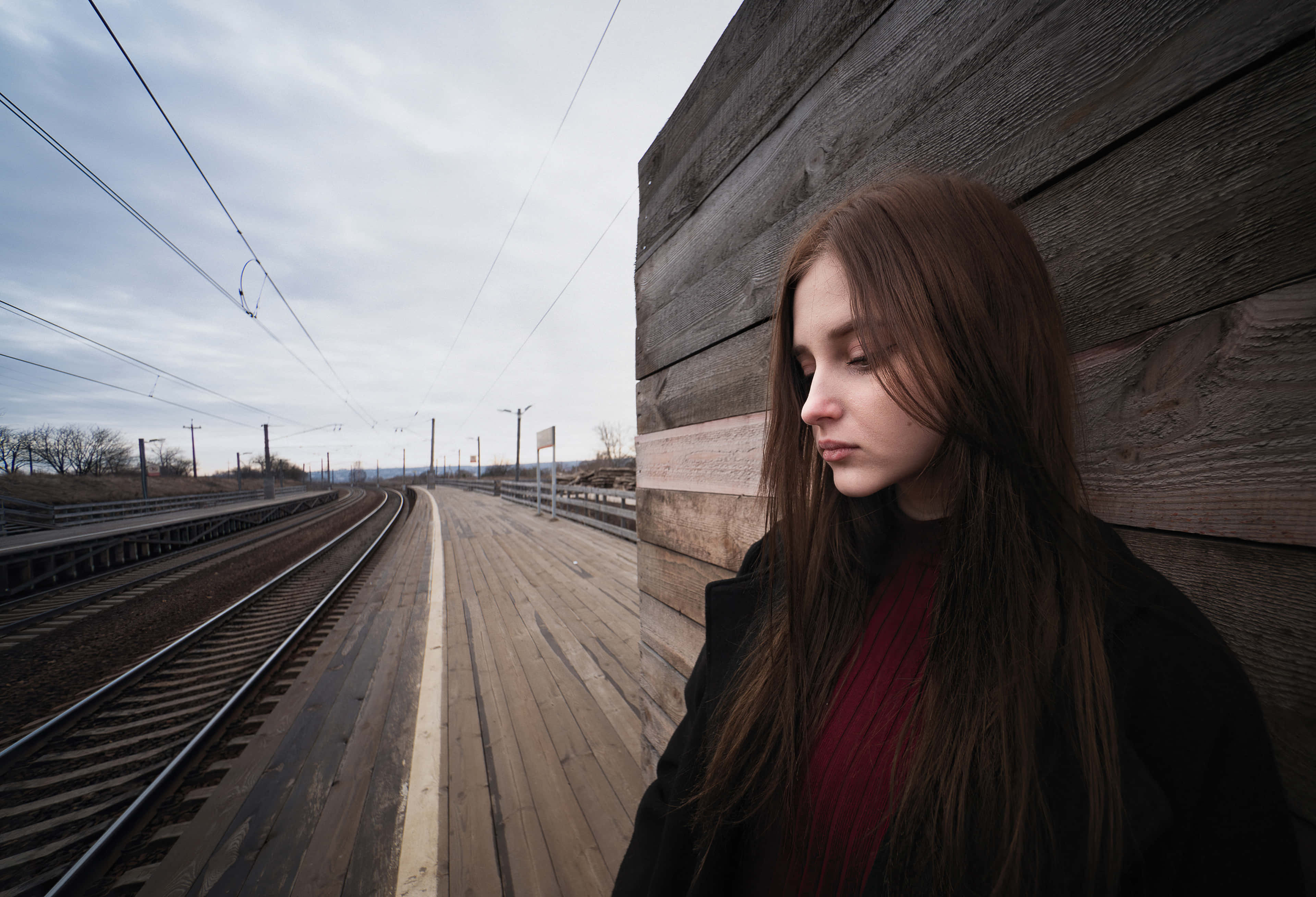 teenager 桌布 壁紙 — 一位長髮棕色的年輕女子靠著風化的木製火車月台牆壁，若有所思地凝視著空蕩蕩的鐵軌延伸至灰濛濛的遠方。