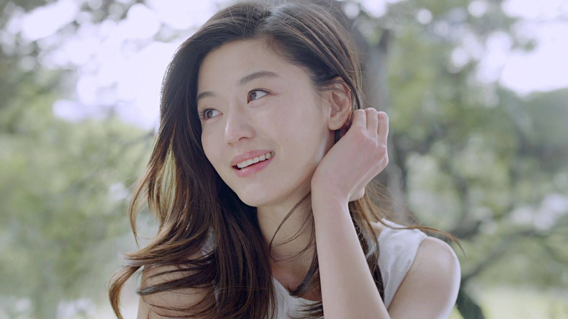 Jun Ji Hyun 桌布 壁紙 — 一位長髮棕色的年輕女性，在柔和的模糊綠色背景前，輕觸耳朵，溫柔微笑。