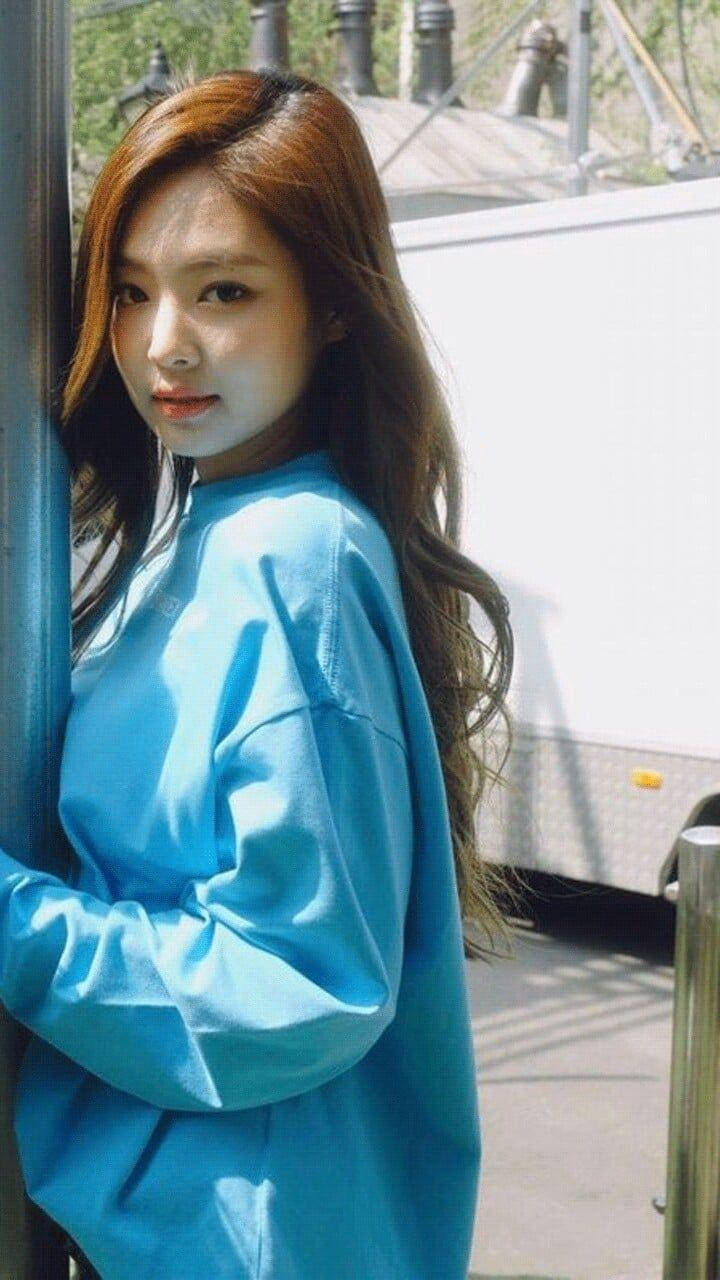 Jennie 桌布 壁紙 — 一位長髮棕色的年輕女性穿著亮藍色寬鬆襯衫，在戶外靠著柱子。
