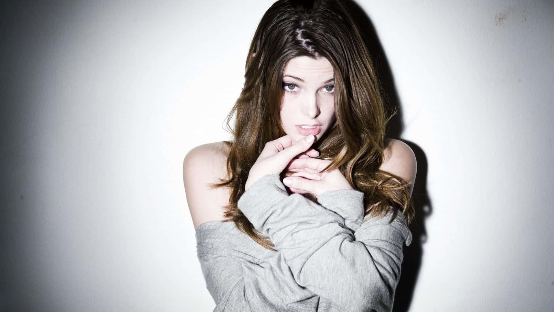 ashley greene 桌布 壁紙 — 一位長髮棕色的年輕女性穿著灰色露肩毛衣，雙手靠近下巴，在純白牆壁前擺姿勢。
