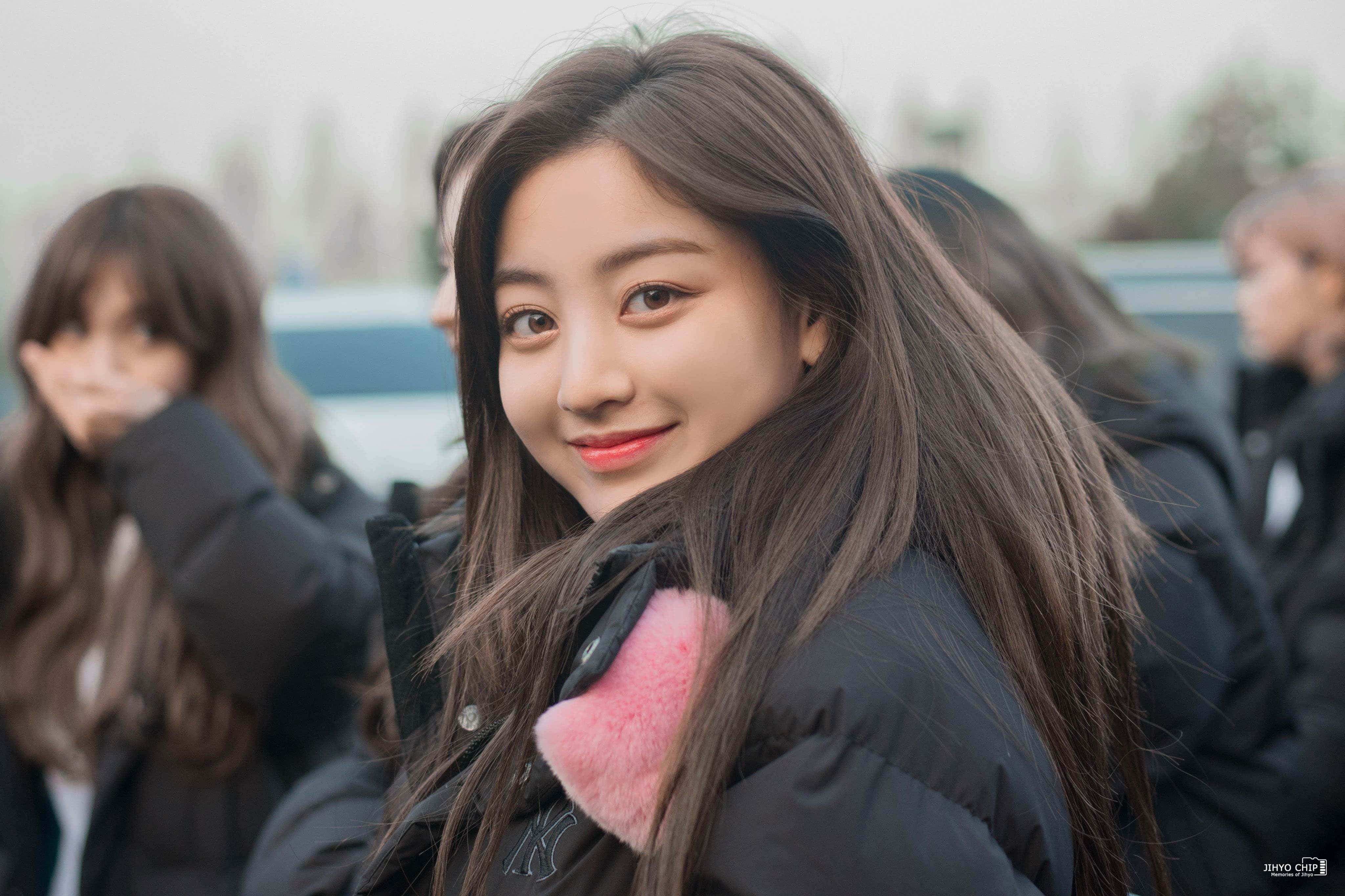 twice 桌布 壁紙 — 一位長髮棕色的年輕女性穿著黑色羽絨外套，搭配粉紅色毛絨內襯的連帽，露出溫暖的笑容。
