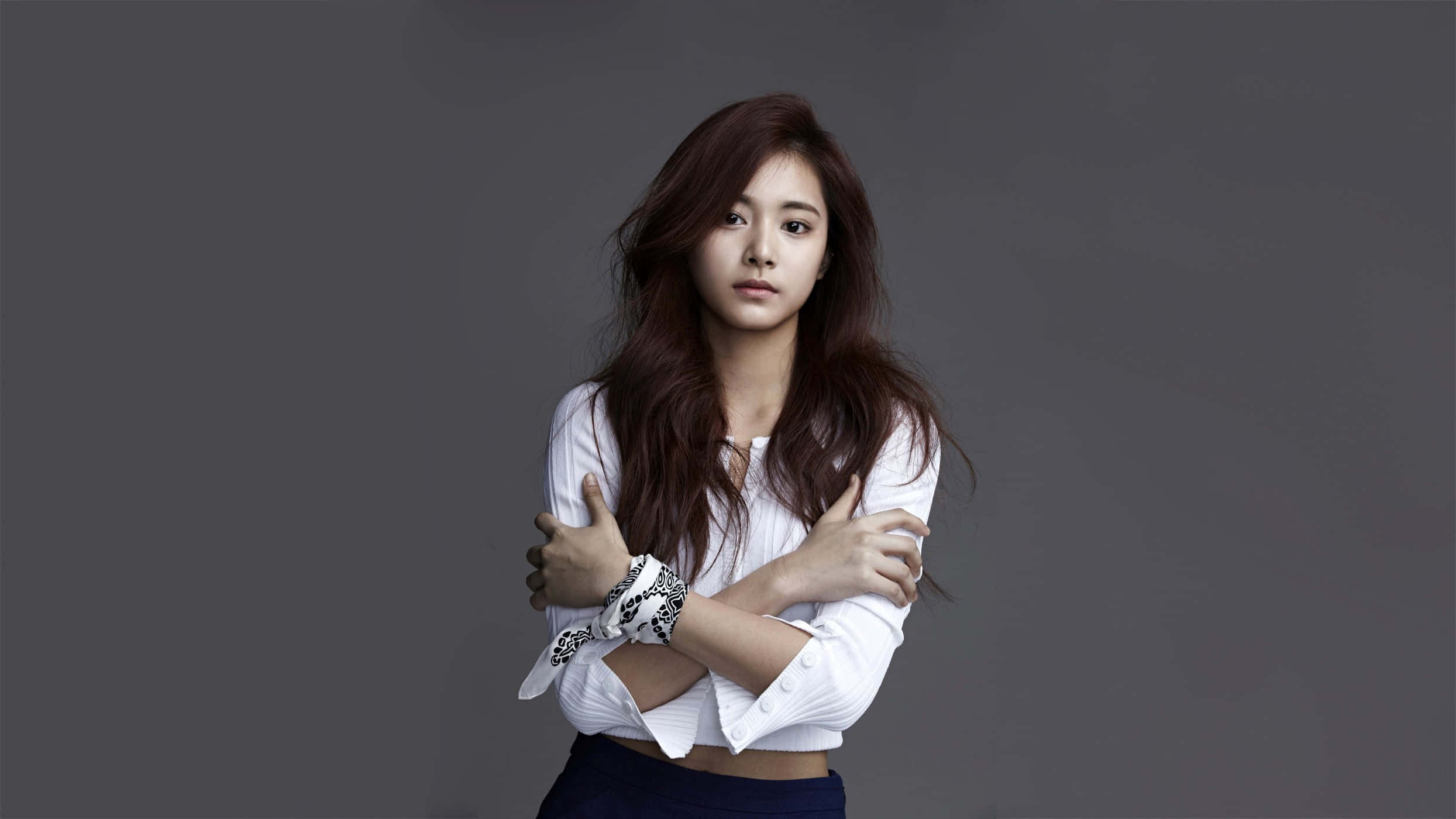 twice 桌布 壁紙 — 一位長髮棕色的年輕女性站在灰色背景前，雙臂交叉在胸前，穿著白色短上衣和深色褲子。