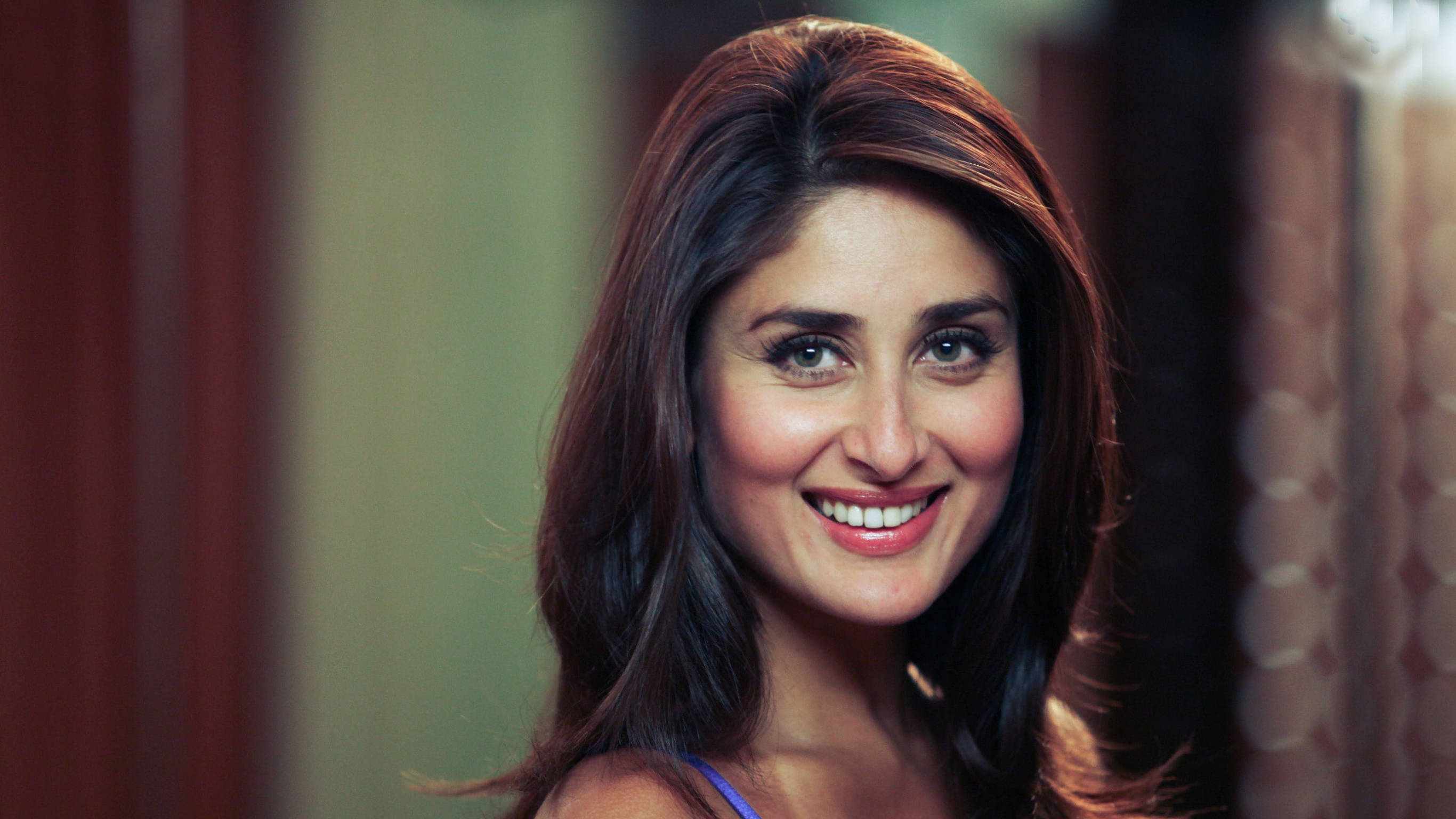 Kareena 桌布 壁紙 — 一位長髮深色、光彩照人的女性在柔和光線的室內環境中對著鏡頭溫暖微笑。