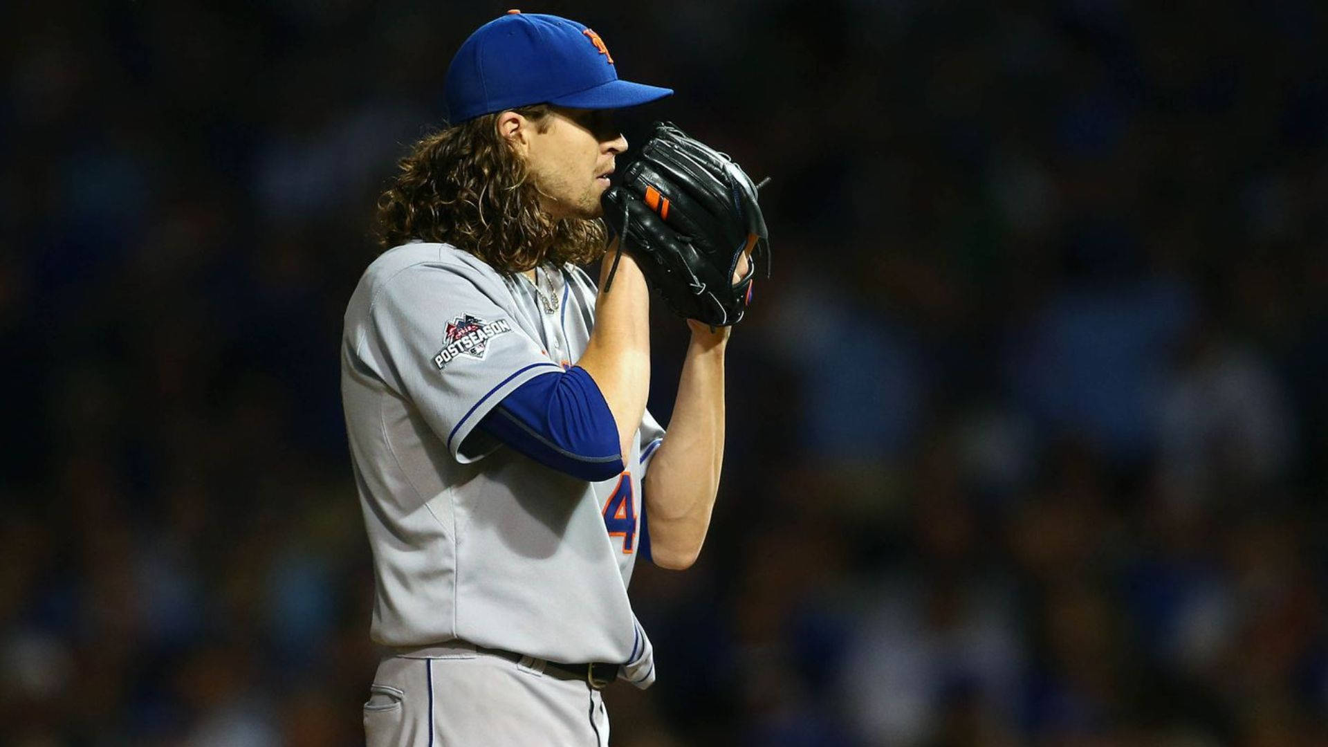 Degrom 桌布 壁紙 — 一位長髮的棒球投手，戴著藍色帽子、穿著灰色制服，在夜間球場上將手套貼在臉上。