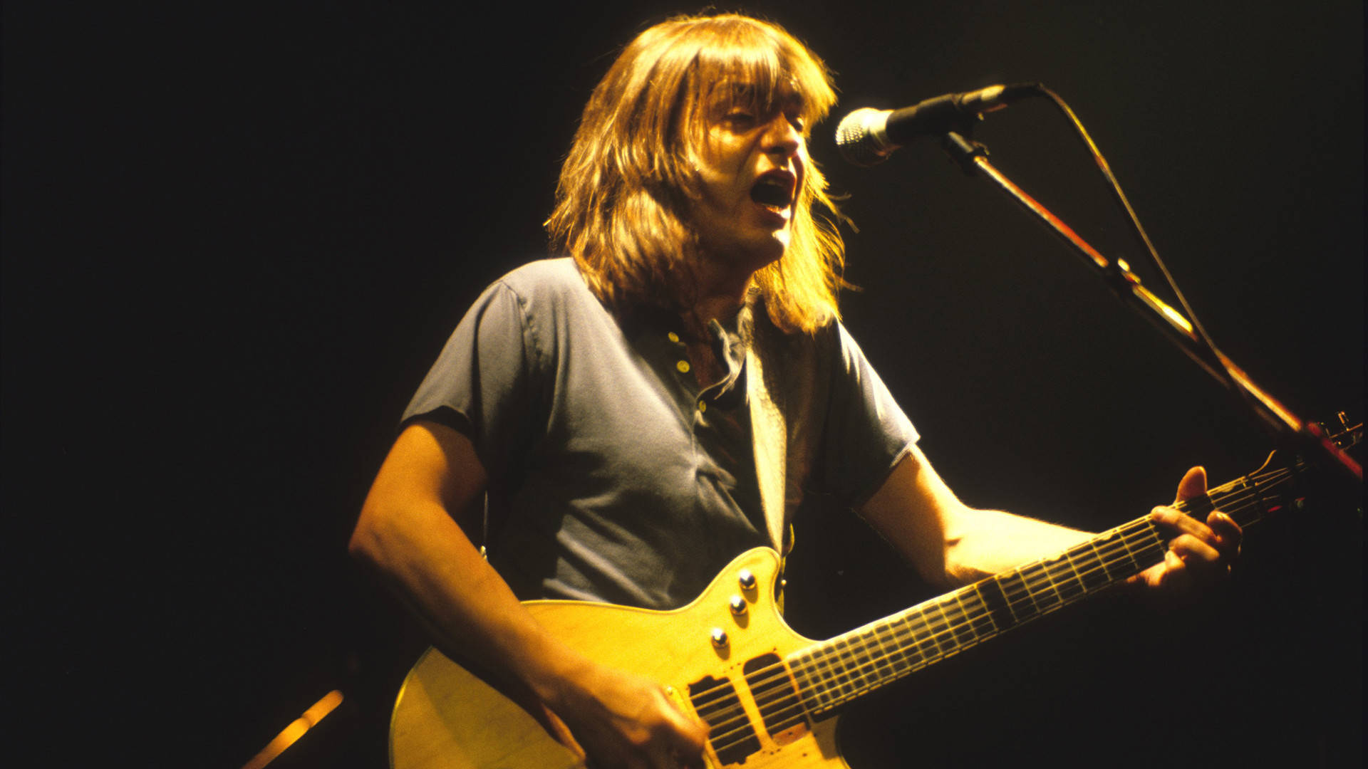Van Halen 桌布 壁紙 — 一位長髮音樂家在黑暗背景下，於溫暖的舞台燈光下熱情地彈奏淺色電吉他。