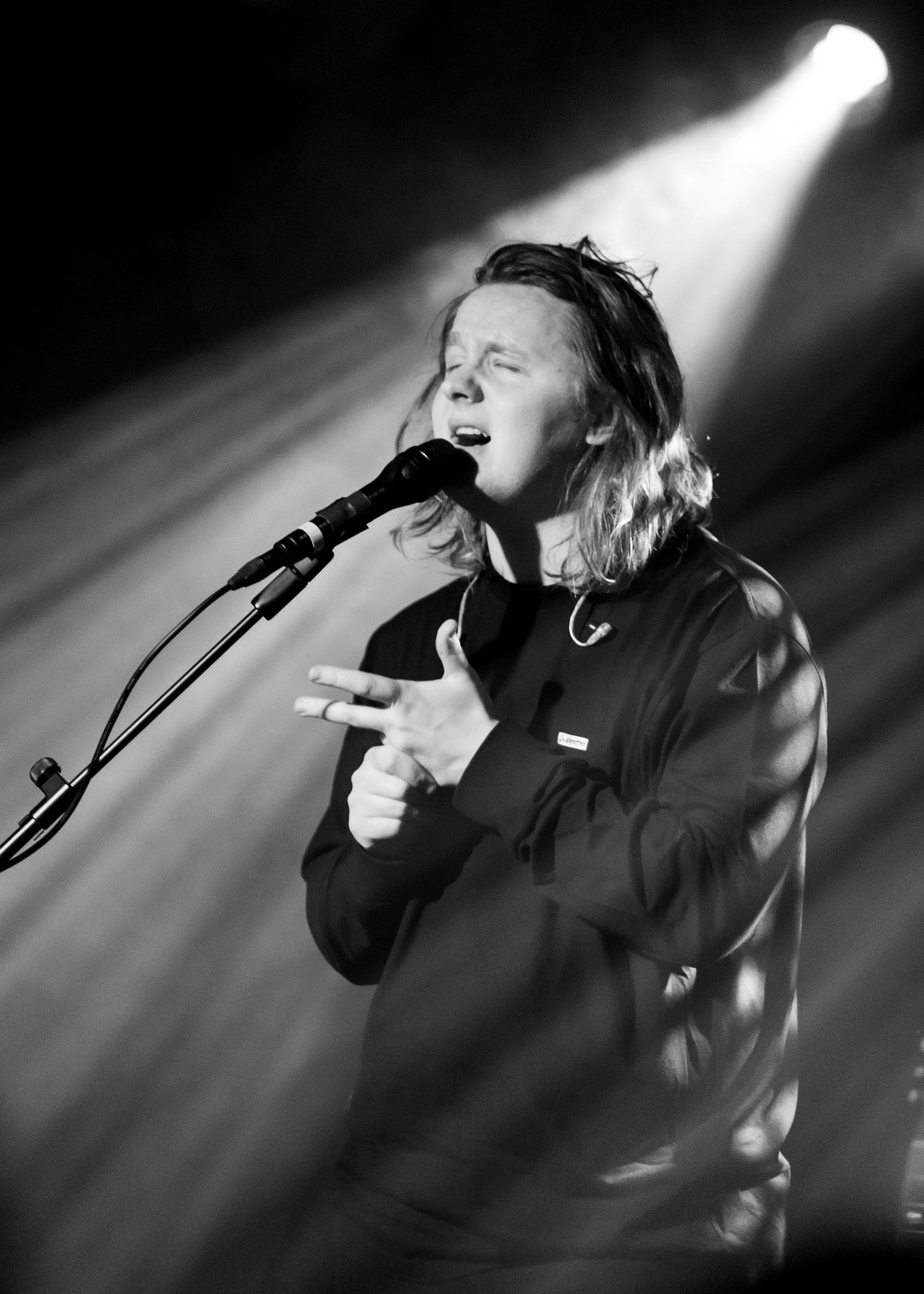 Lewis Capaldi 桌布 壁紙 — 一位長髮音樂家在黑暗舞台上的戲劇性聚光燈下，熱情地對著麥克風唱歌。