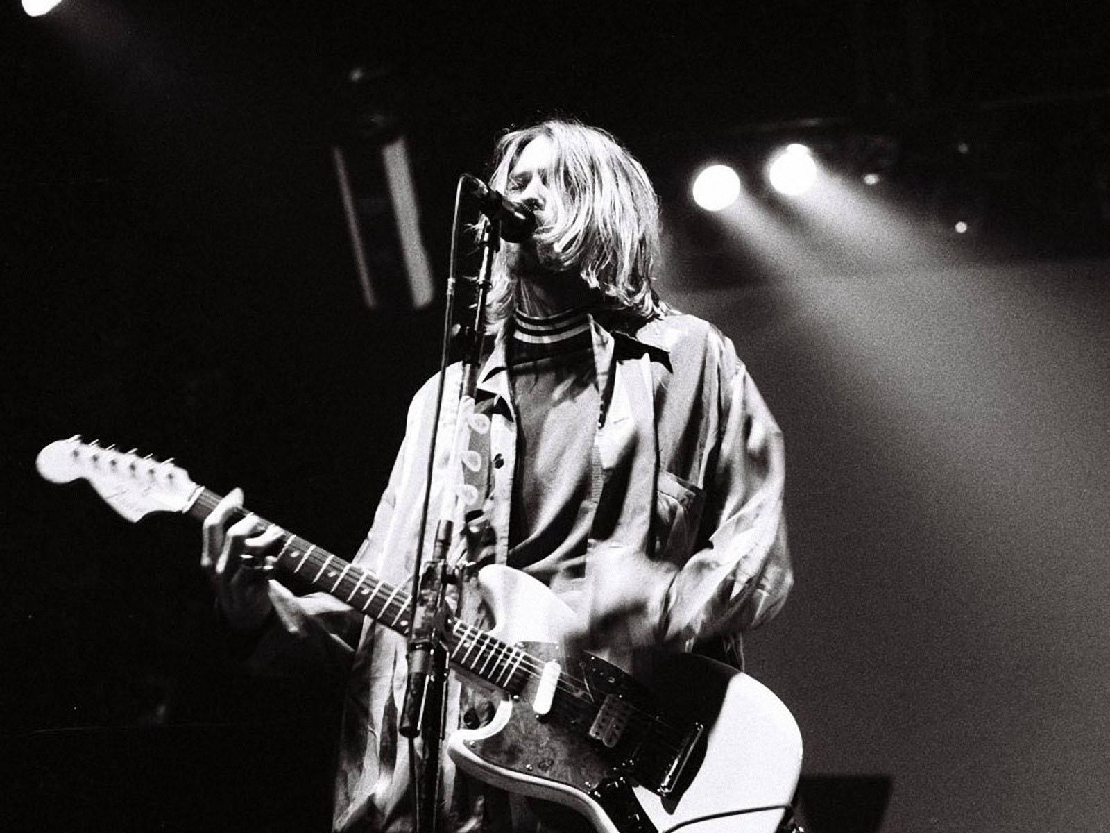 nirvana 4K 桌布 壁紙 — 一位長髮音樂家在黑白明亮的舞台燈光下彈奏電吉他並對著麥克風唱歌。