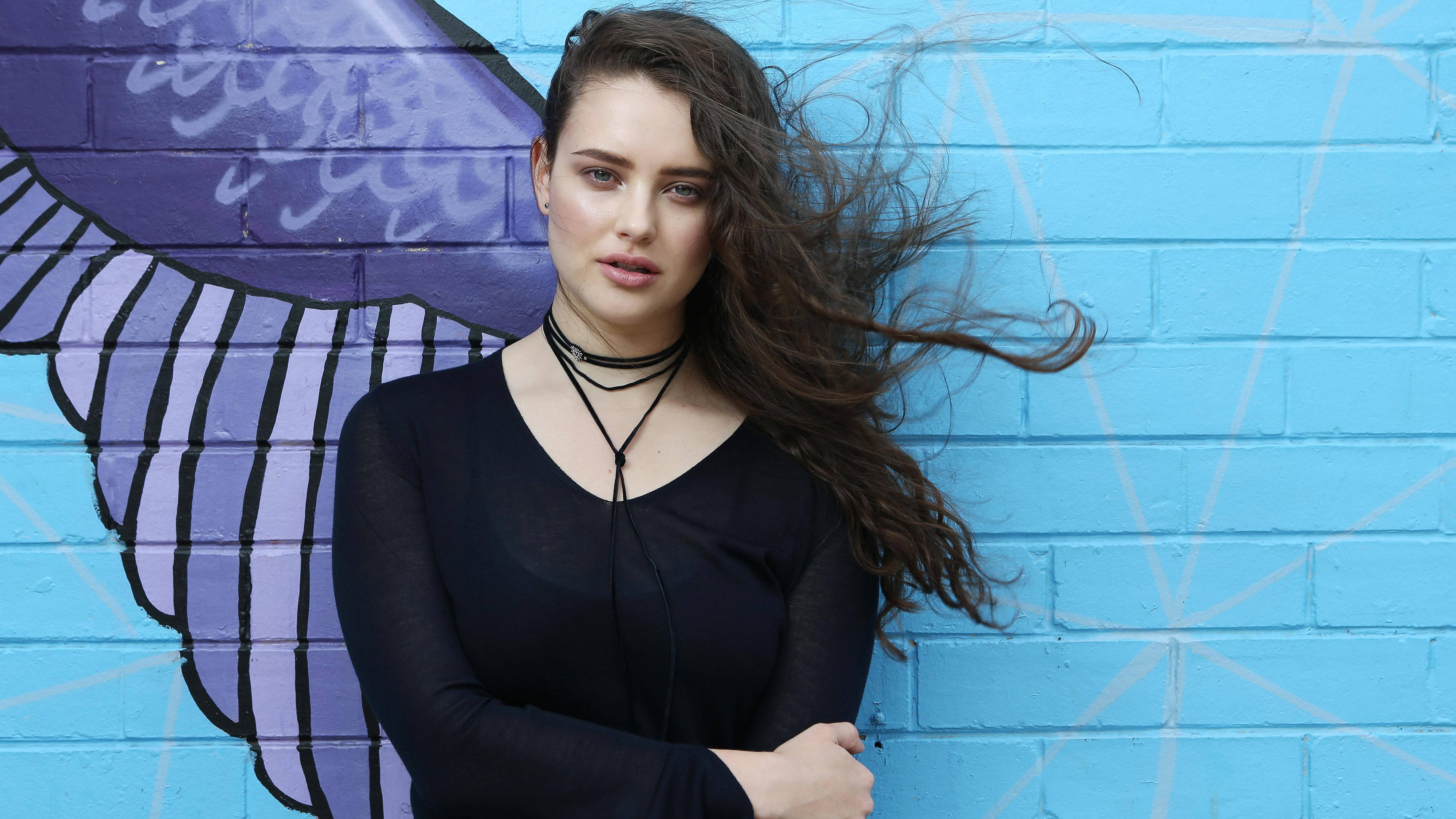 Katherine Langford 桌布 壁紙 — 一位長髮飄逸的年輕女子站在藍色磚牆前，牆上裝飾著一幅巨大的紫色蝴蝶壁畫。